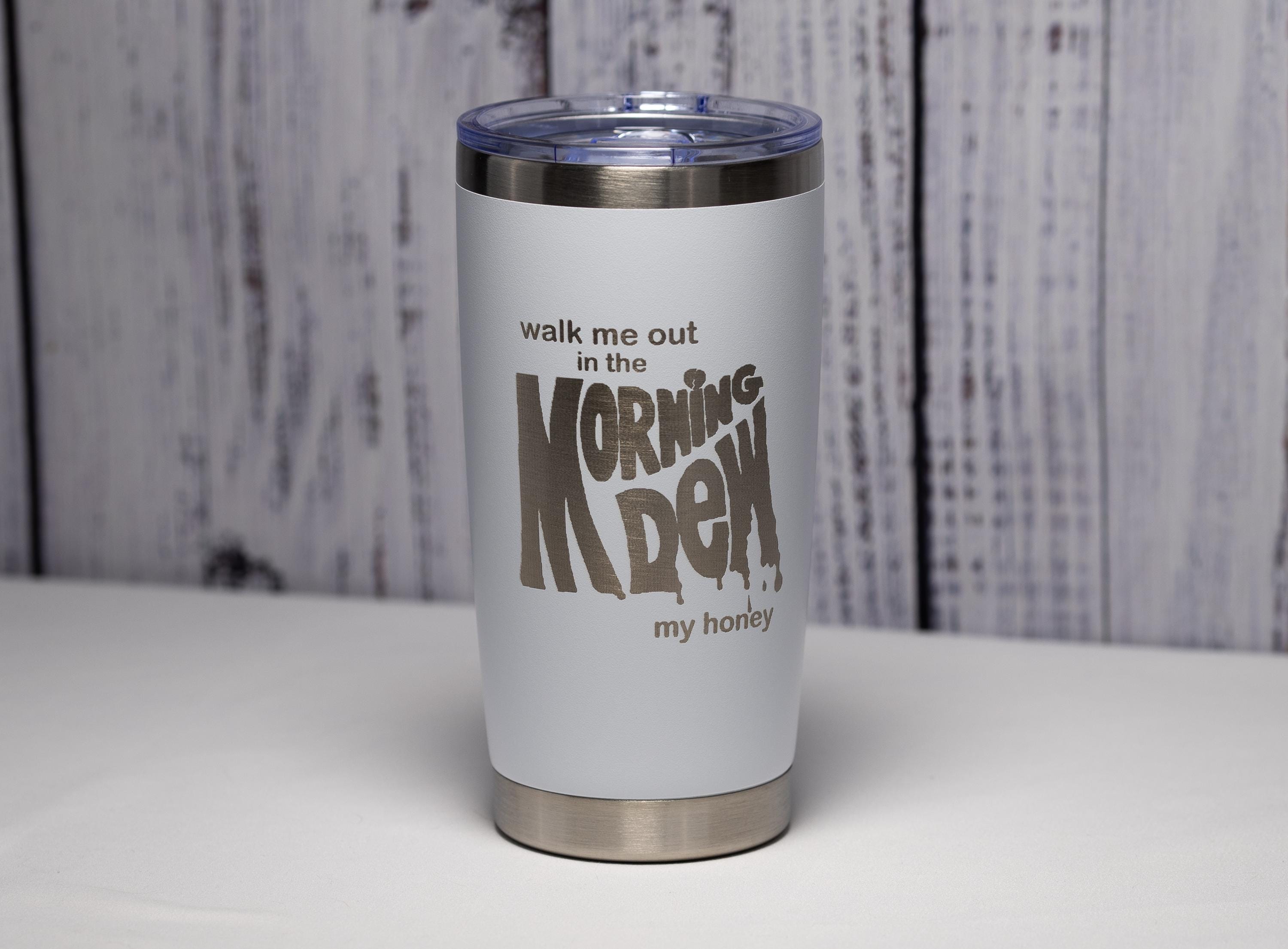May include: A white insulated tumbler with a silver rim and lid. The tumbler features the text "walk me out in the MORNING DEW my honey" in a brown, stylized font. The tumbler is designed to keep beverages hot or cold.
