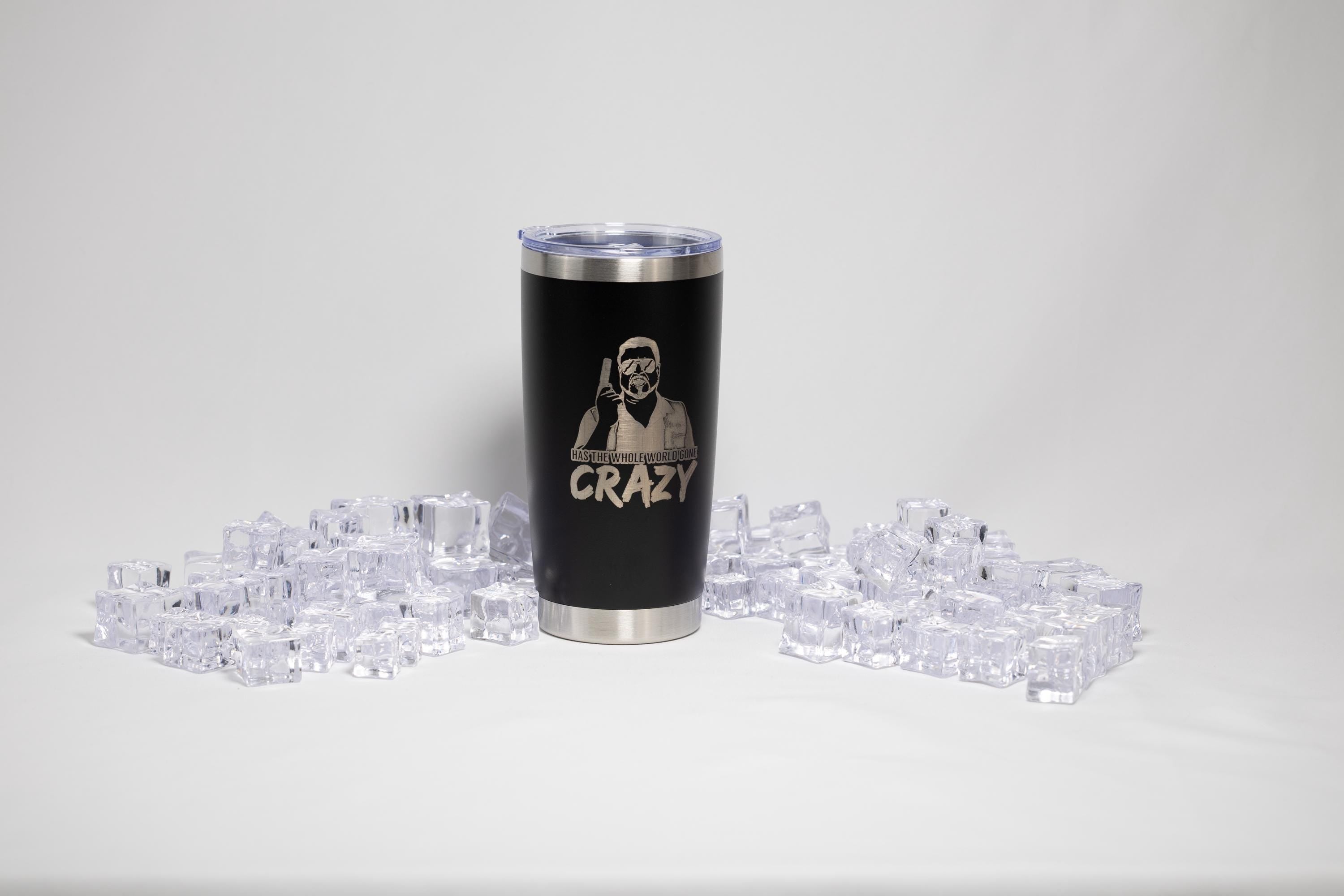 A black tumbler with a silver lid, featuring a printed design, is surrounded by numerous clear ice cubes on a white background.