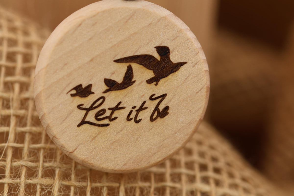 May include: A wooden round pendant with a laser-etched design. The design features three birds in flight above the words 'Let it Be' in a handwritten font. The pendant is light brown with a natural wood grain appearance. The background is a blurred, neutral tone.