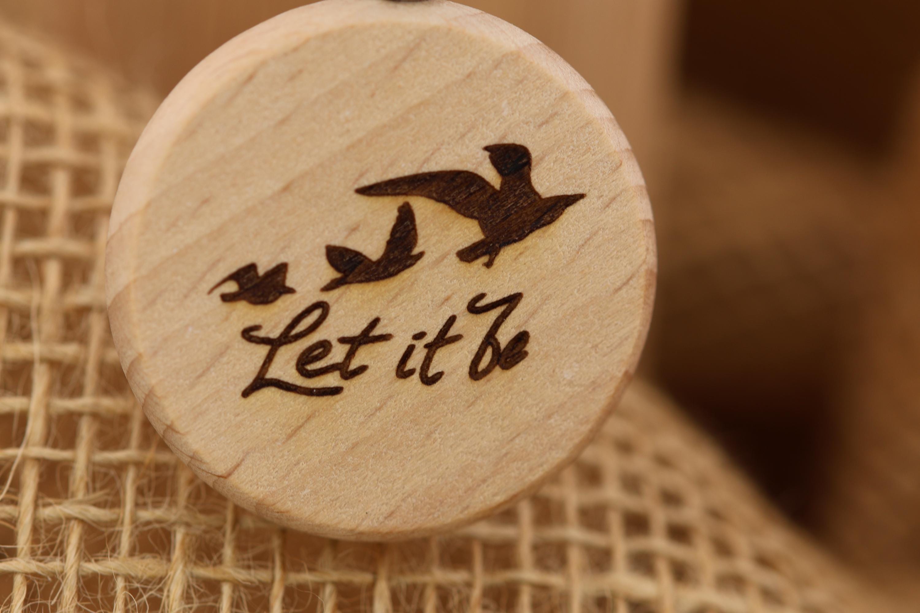 May include: A wooden round pendant with a laser-etched design. The design features three birds in flight above the words 'Let it Be' in a handwritten font. The pendant is light brown with a natural wood grain appearance. The background is a blurred, neutral tone.
