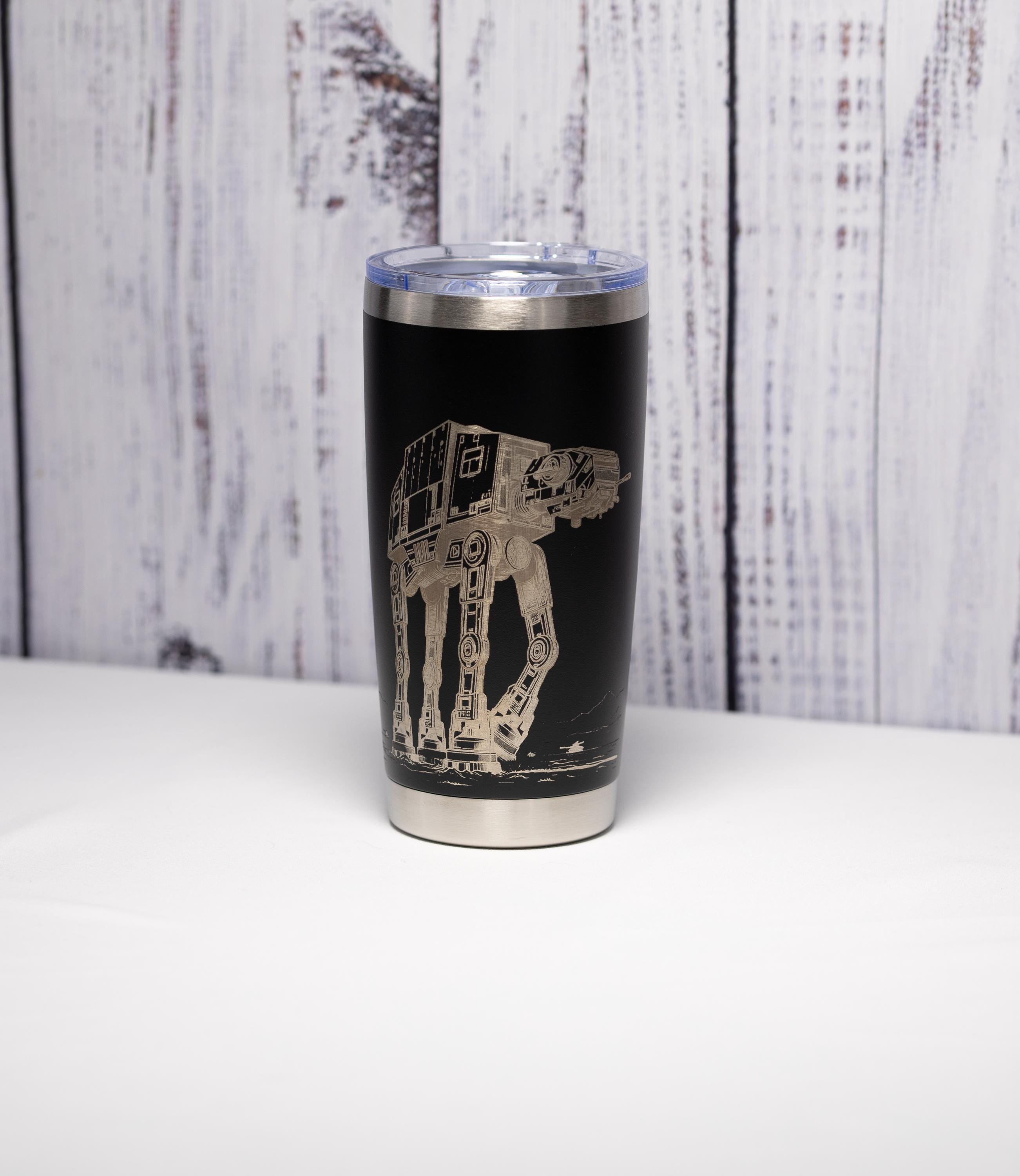 May include: Black stainless steel tumbler with a clear lid. The tumbler features a detailed etched design of an AT-AT walker from Star Wars. The design is in a light silver color. The tumbler is approximately 7 inches tall.