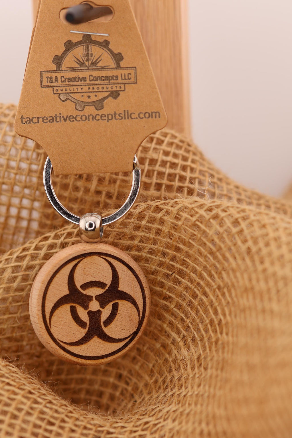 May include: A wooden keychain with a circular pendant featuring a laser-engraved biohazard symbol. The keychain has a silver ring and a brown tag with the text 'T&A Creative Concepts LLC QUALITY PRODUCTS tacreativeconceptsllc.com'.