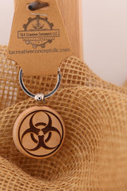 May include: A wooden keychain with a circular pendant featuring a laser-engraved biohazard symbol. The keychain has a silver ring and a brown tag with the text 'T&A Creative Concepts LLC QUALITY PRODUCTS tacreativeconceptsllc.com'.
