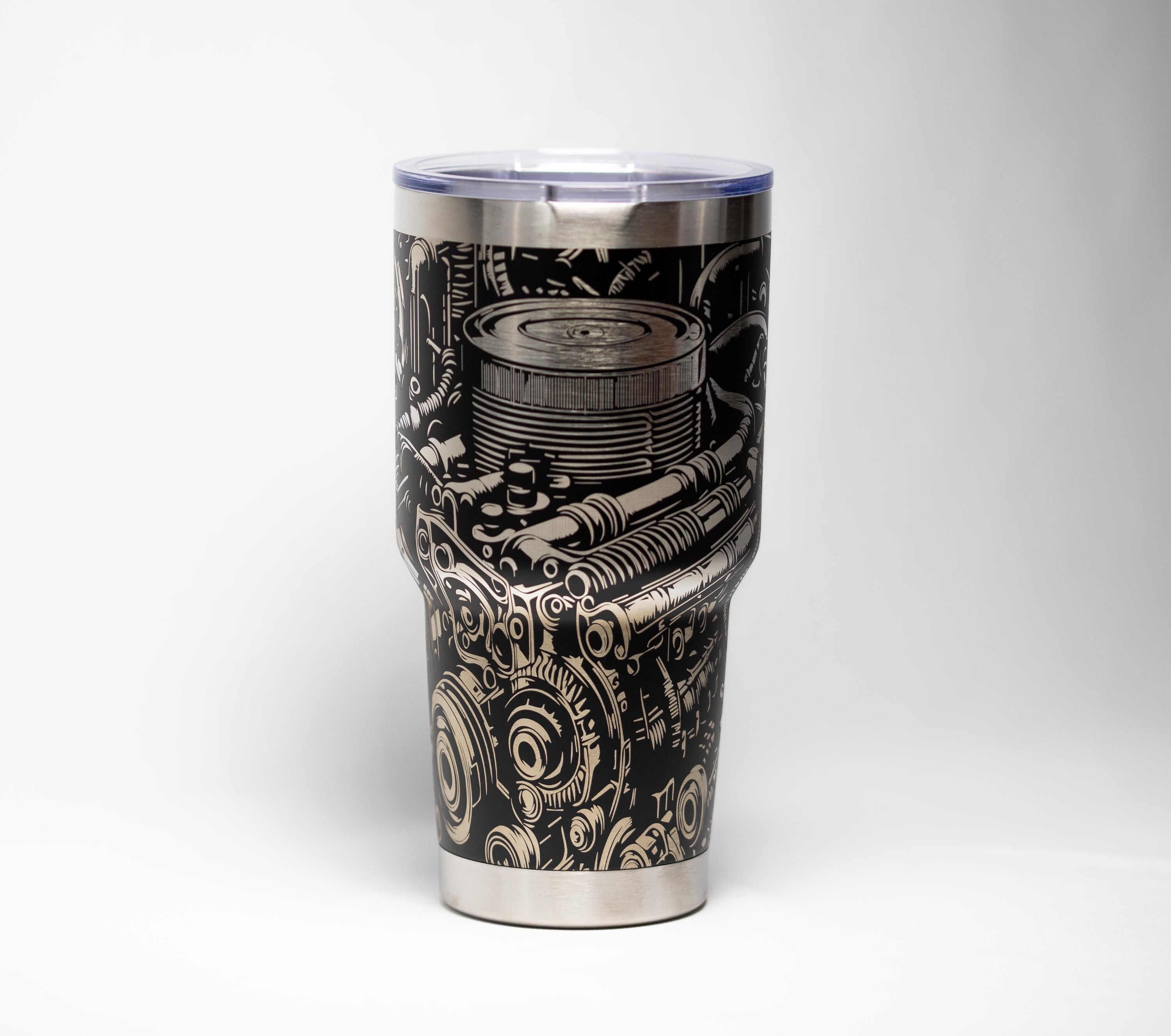 May include: A stainless steel tumbler with a black and silver engraved design of an engine's internal components. The tumbler has a clear plastic lid and a silver rim and base. The design features gears, pipes, and other mechanical elements.