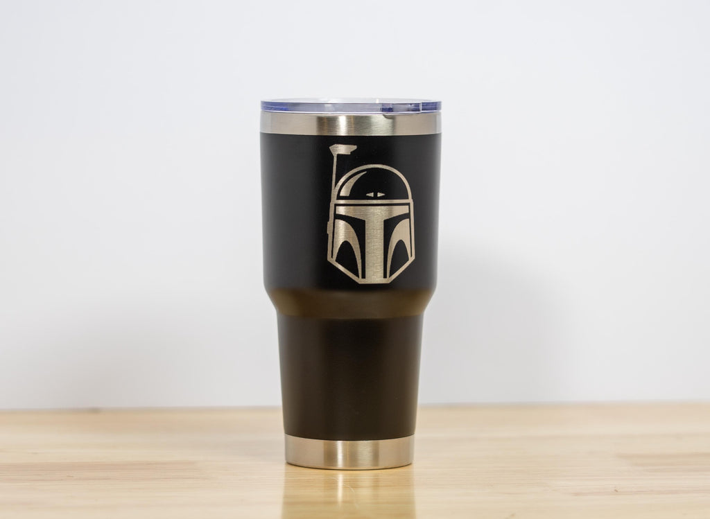 May include: A black insulated tumbler with a silver rim and lid. The tumbler features a gold-colored graphic of a Mandalorian helmet. The tumbler is designed to keep beverages hot or cold. The tumbler is suitable for travel and everyday use.