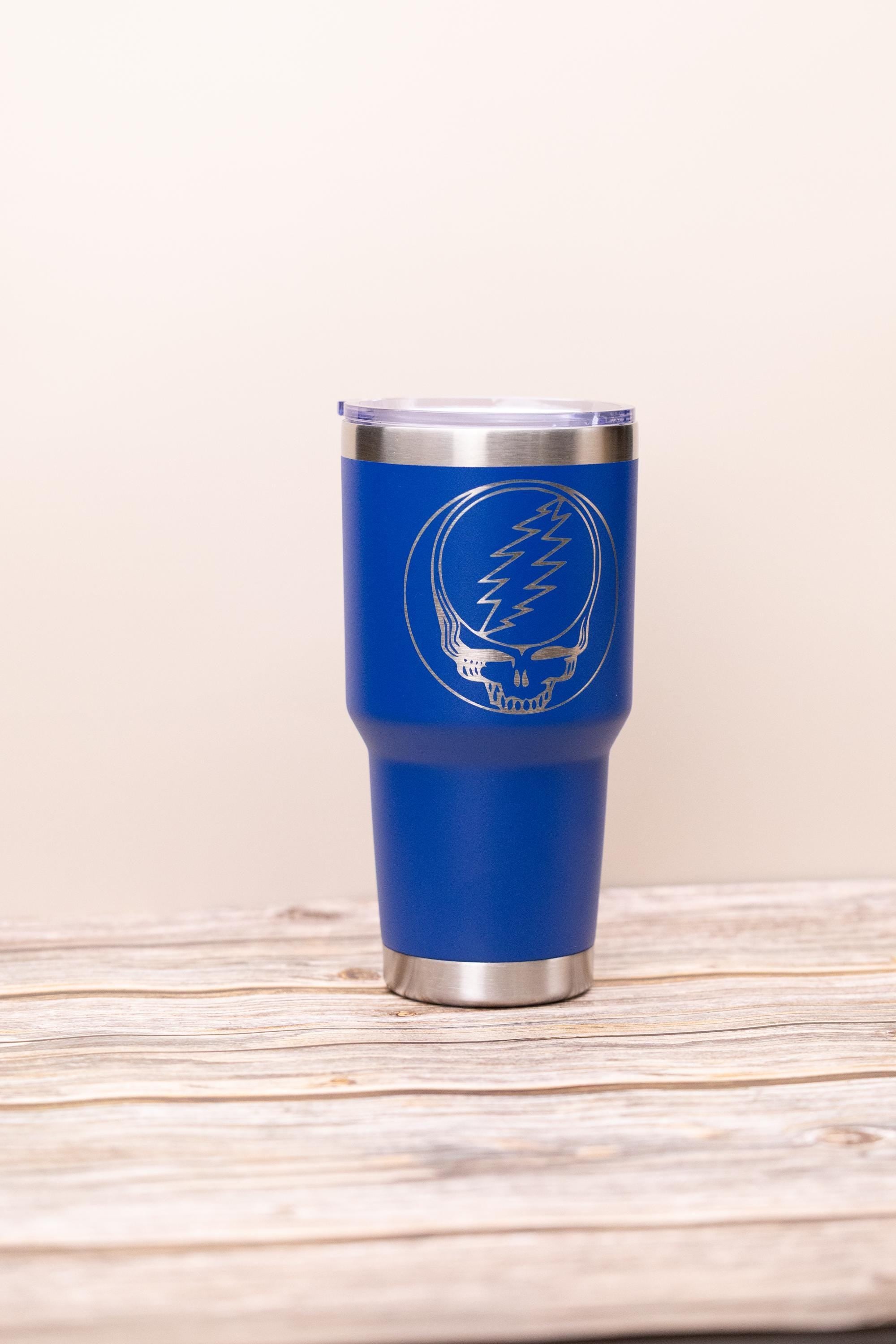 May include: A blue stainless steel tumbler with a silver rim and lid. The tumbler features a silver graphic of a skull with a lightning bolt on the front. The tumbler is sitting on a wooden surface.