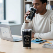 A man is sitting at a desk, drinking from a black tumbler with the text "SCIENCE DOESN'T CARE WHAT YOU BELIEVE" printed on it. The desk also has a laptop, a notebook, and some books.