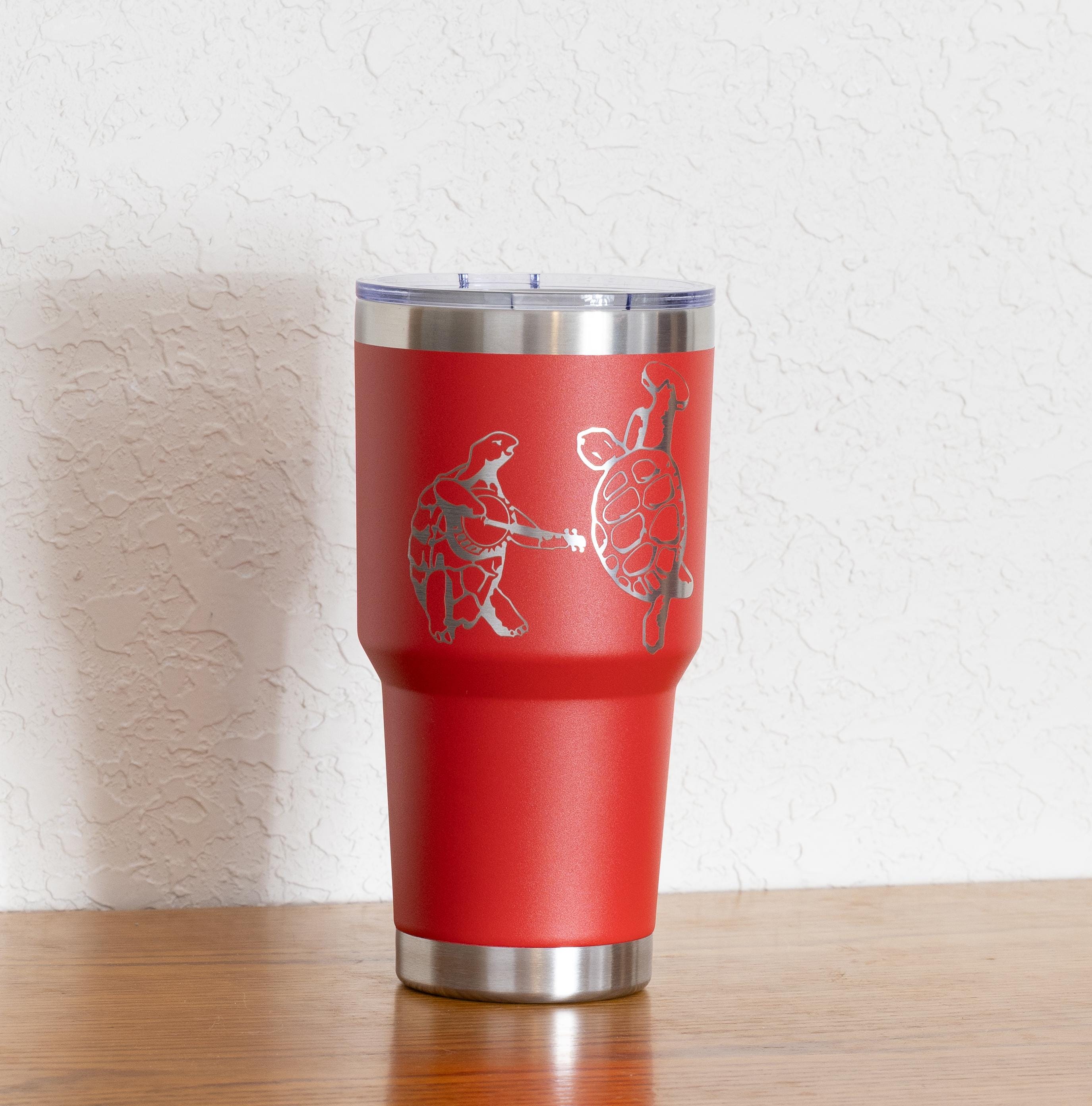 May include: A red insulated tumbler with a silver lid and base. The tumbler features a silver engraving of two turtles, one holding a guitar. The tumbler is designed to keep beverages cold or hot.