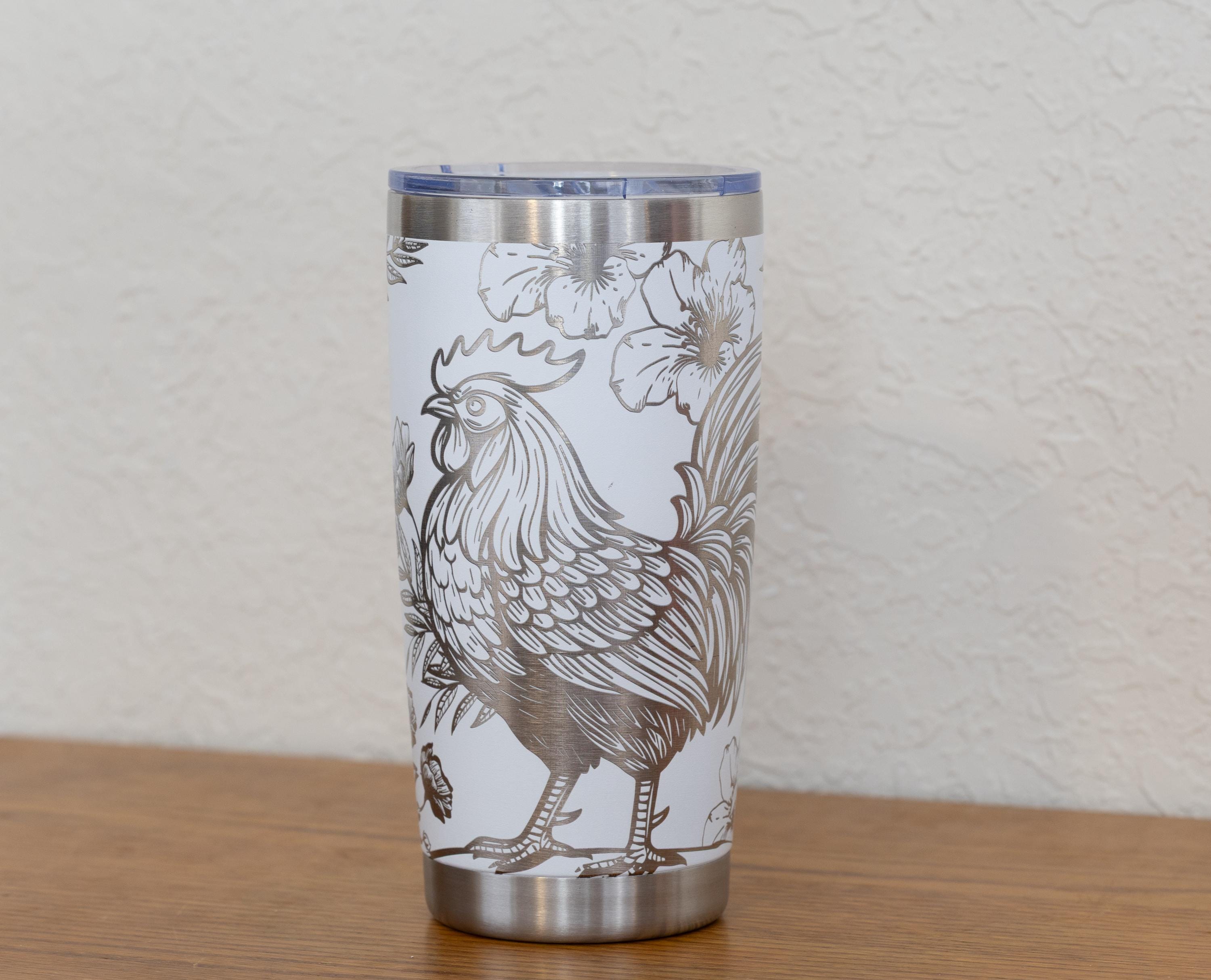 May include: A stainless steel tumbler with a white background and a detailed silver rooster illustration. The design includes floral accents. The tumbler has a silver rim and a clear lid.