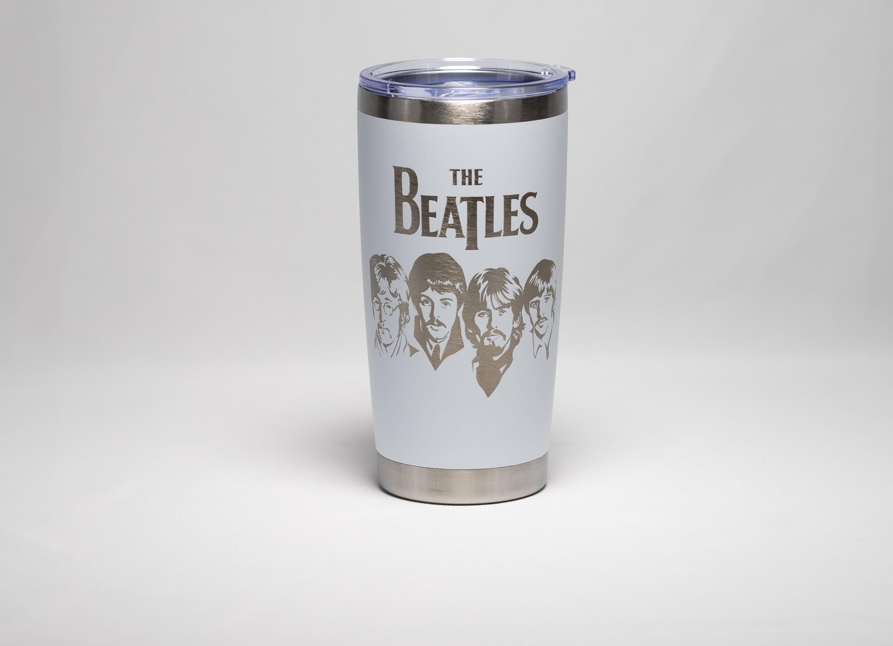 May include: A white insulated tumbler featuring the band name "THE BEATLES" in a bold font, with etched portraits of the band members below. The tumbler has a silver rim and base, and a clear plastic lid.