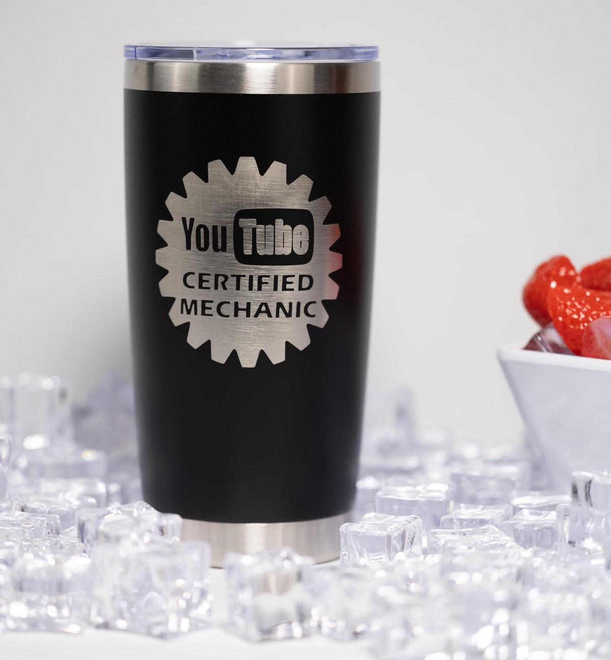 A black tumbler with a silver lid, featuring a logo that says "YouTube Certified Mechanic". The tumbler is placed on a surface with ice cubes, and there is a bowl of strawberries in the background.