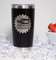 A black tumbler with a silver lid, featuring a logo that says "YouTube Certified Mechanic". The tumbler is placed on a surface with ice cubes, and there is a bowl of strawberries in the background.