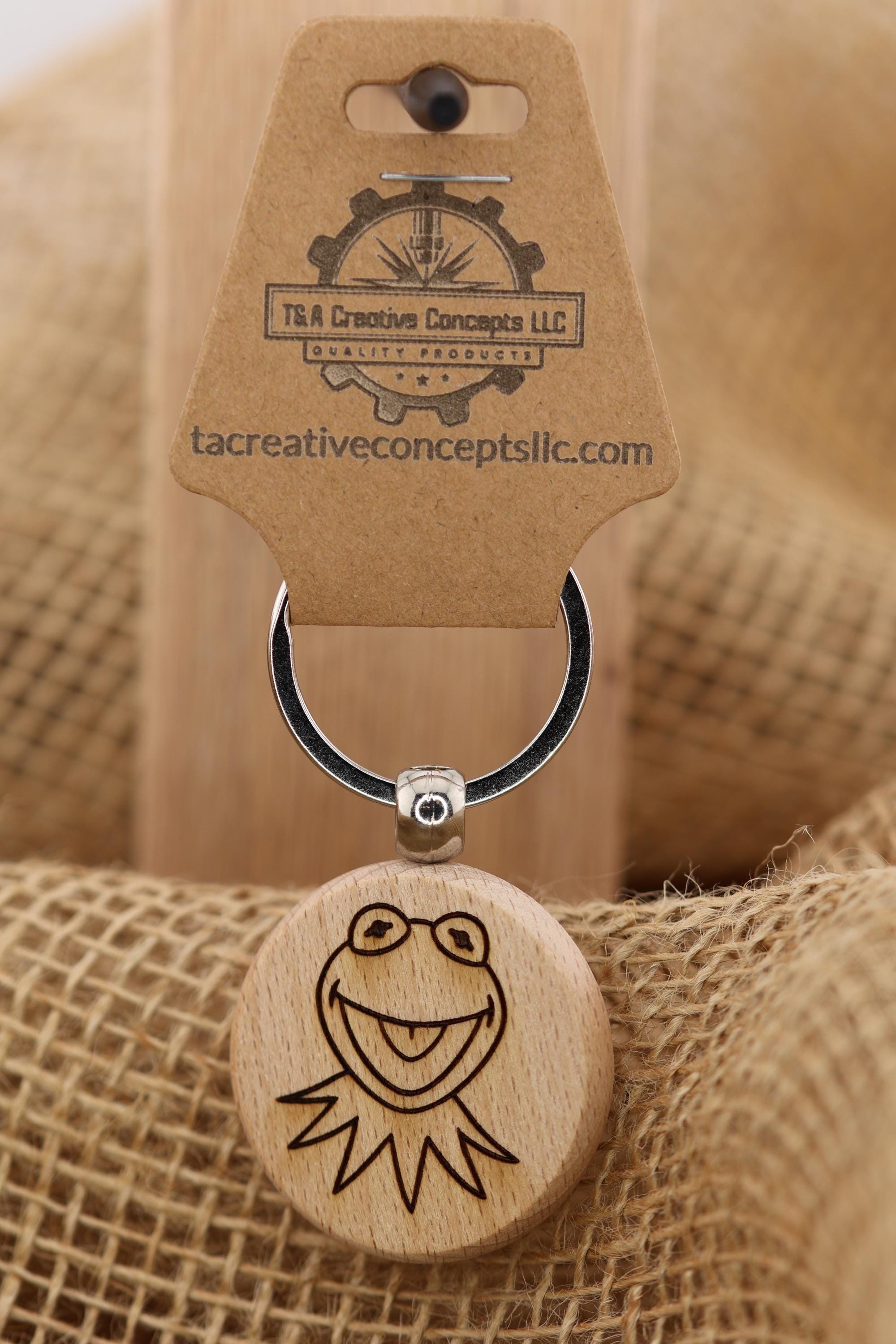 May include: A wooden Kermit the Frog keychain. The round charm features a laser-etched image of Kermit's face with his signature smile. The keychain includes a metal ring and a tag with the text 'T&A Creative Concepts LLC Quality Products' and the website address. The background is a burlap texture.