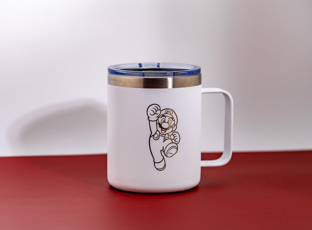 May include: White travel mug with a gold rim and a blue lid. The mug features a gold outline of the character Mario from the Super Mario Bros. video game series. The mug has a handle and is set against a red and white background.
