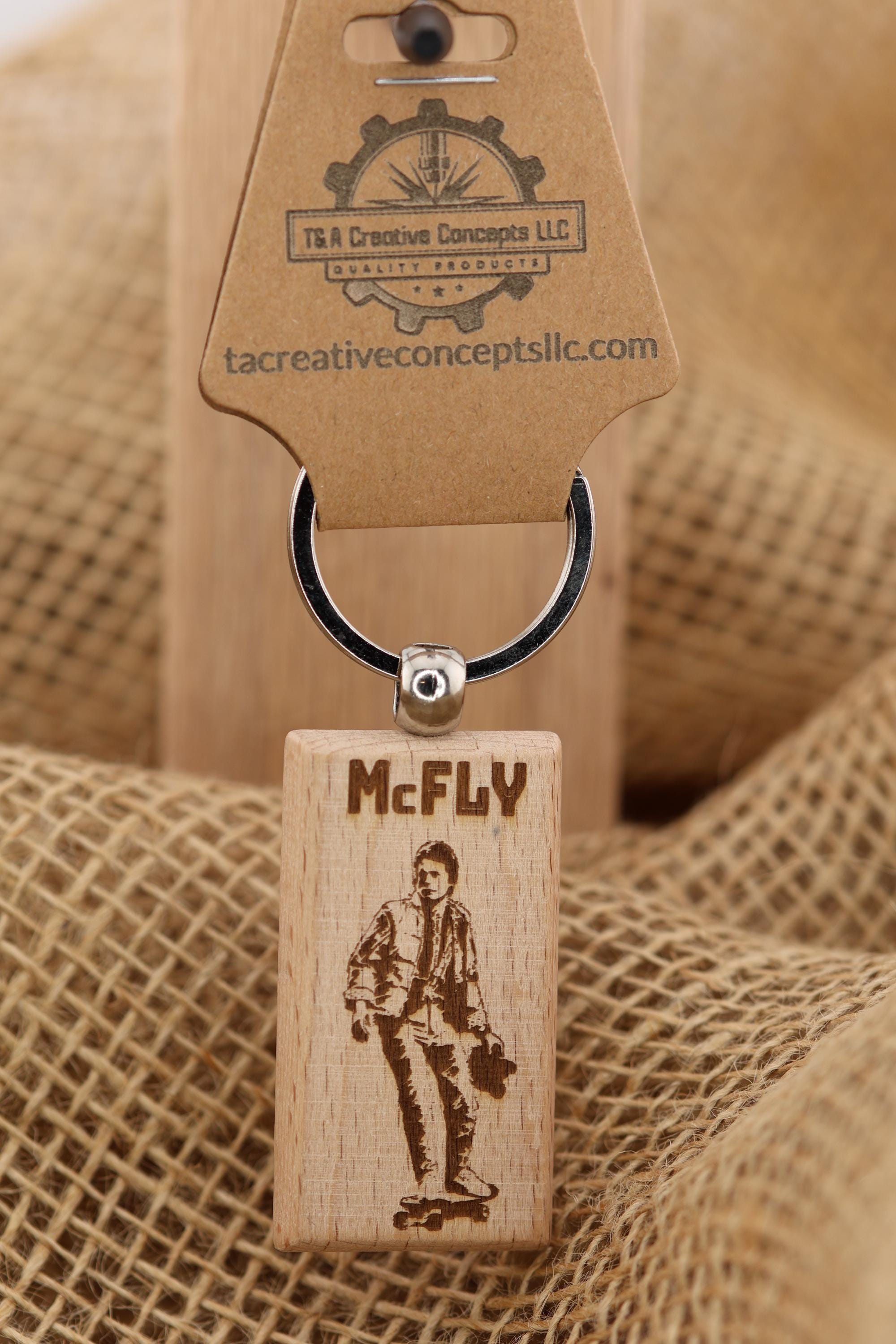 May include: A wooden keychain with the name "McFLY" engraved on it, along with an image of a person on a skateboard. The keychain has a silver ring and a brown tag with the logo of "T&A Creative Concepts LLC".