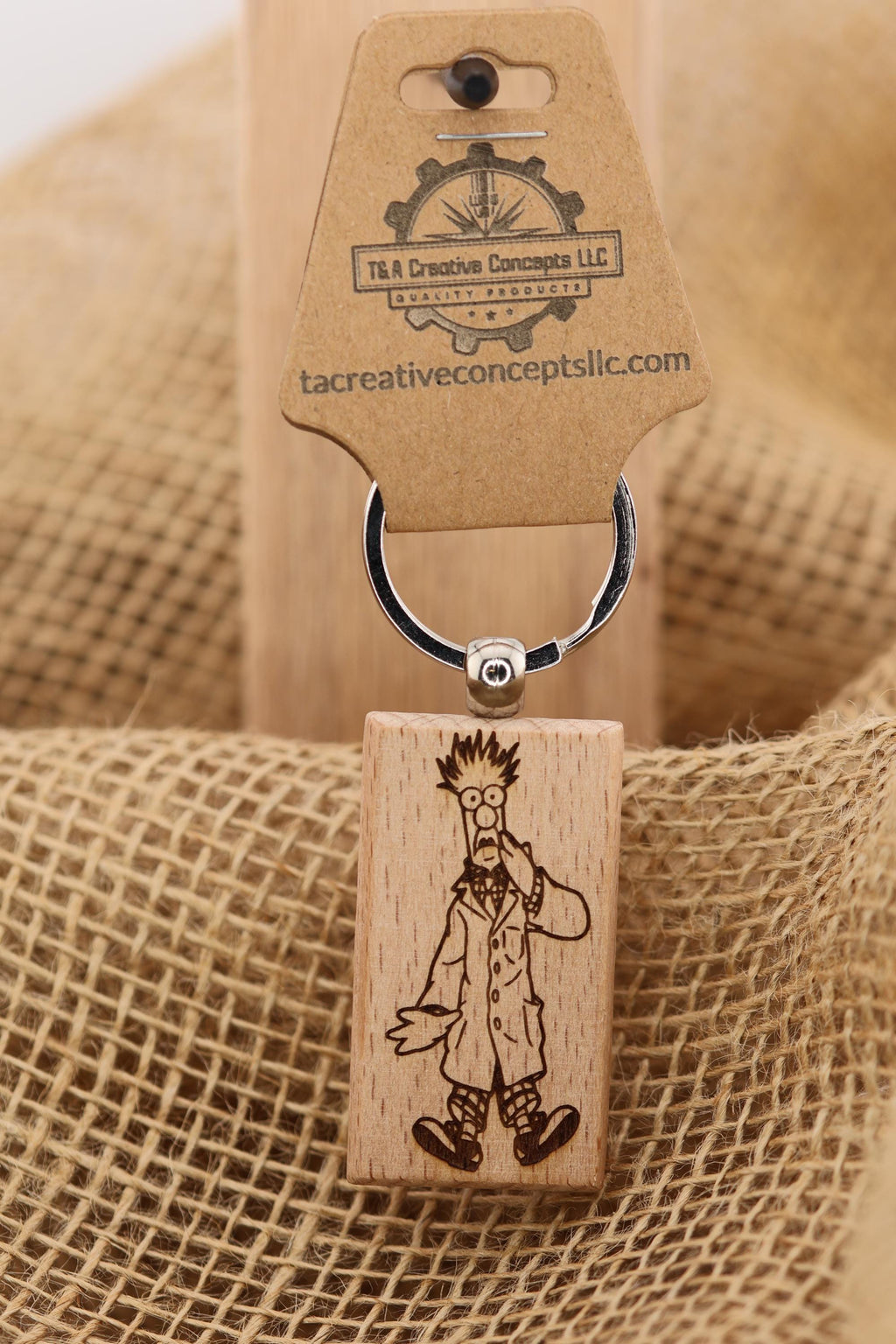 May include: A wooden keychain featuring a cartoon character engraving. The keychain has a rectangular wooden charm with a black and white image of a scientist with glasses and a lab coat. The charm is attached to a metal ring with a brown tag that reads 'T&A Creative Concepts LLC' and 'tacreativeconceptsllc.com'.