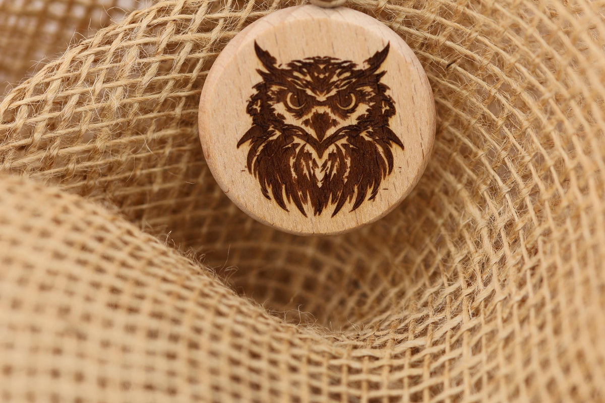 May include: A wooden owl pendant with a detailed engraved design. The pendant is circular and features a brown owl with intricate feather details. The pendant is displayed on a textured, tan burlap background, creating a rustic aesthetic. The owl pendant is a unique piece of jewelry.
