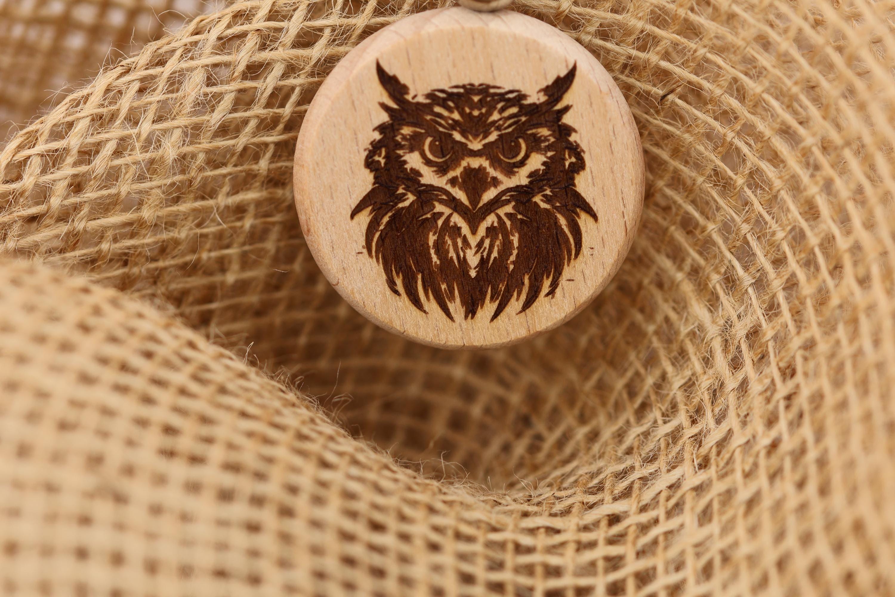May include: A wooden owl pendant with a detailed engraved design. The pendant is circular and features a brown owl with intricate feather details. The pendant is displayed on a textured, tan burlap background, creating a rustic aesthetic. The owl pendant is a unique piece of jewelry.