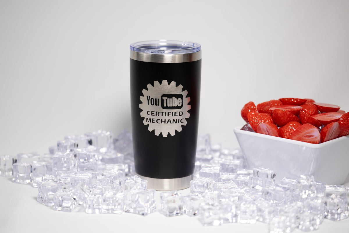 A black tumbler with a YouTube logo and the text "Certified Mechanic" is placed on a bed of ice cubes, with a bowl of red strawberries to the right.