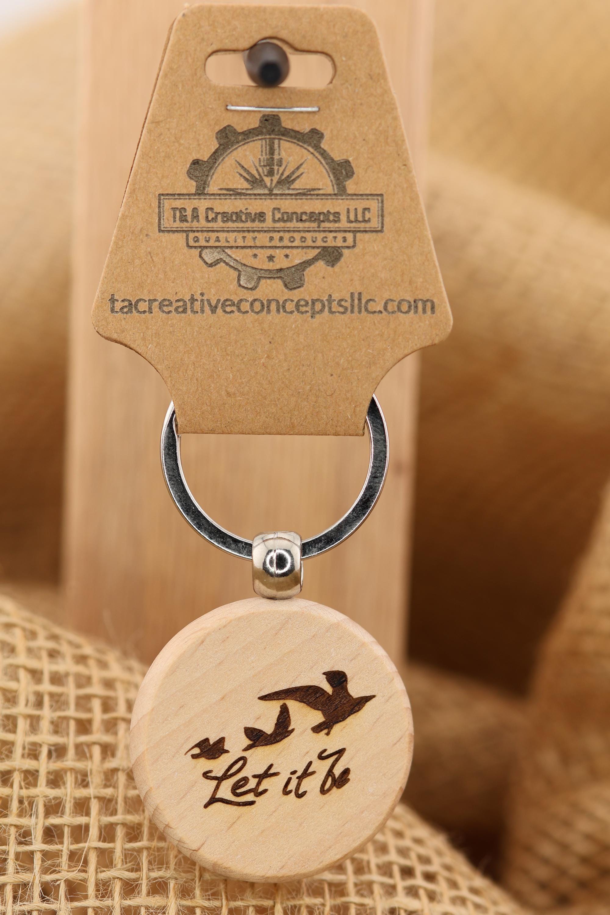 May include: A wooden keychain with a round pendant featuring a laser-etched design of three birds in flight and the words 'Let it be.' The keychain also includes a metal ring and a tag with the text 'T&A Creative Concepts LLC Quality Products' and the website tacreativeconceptsllc.com.