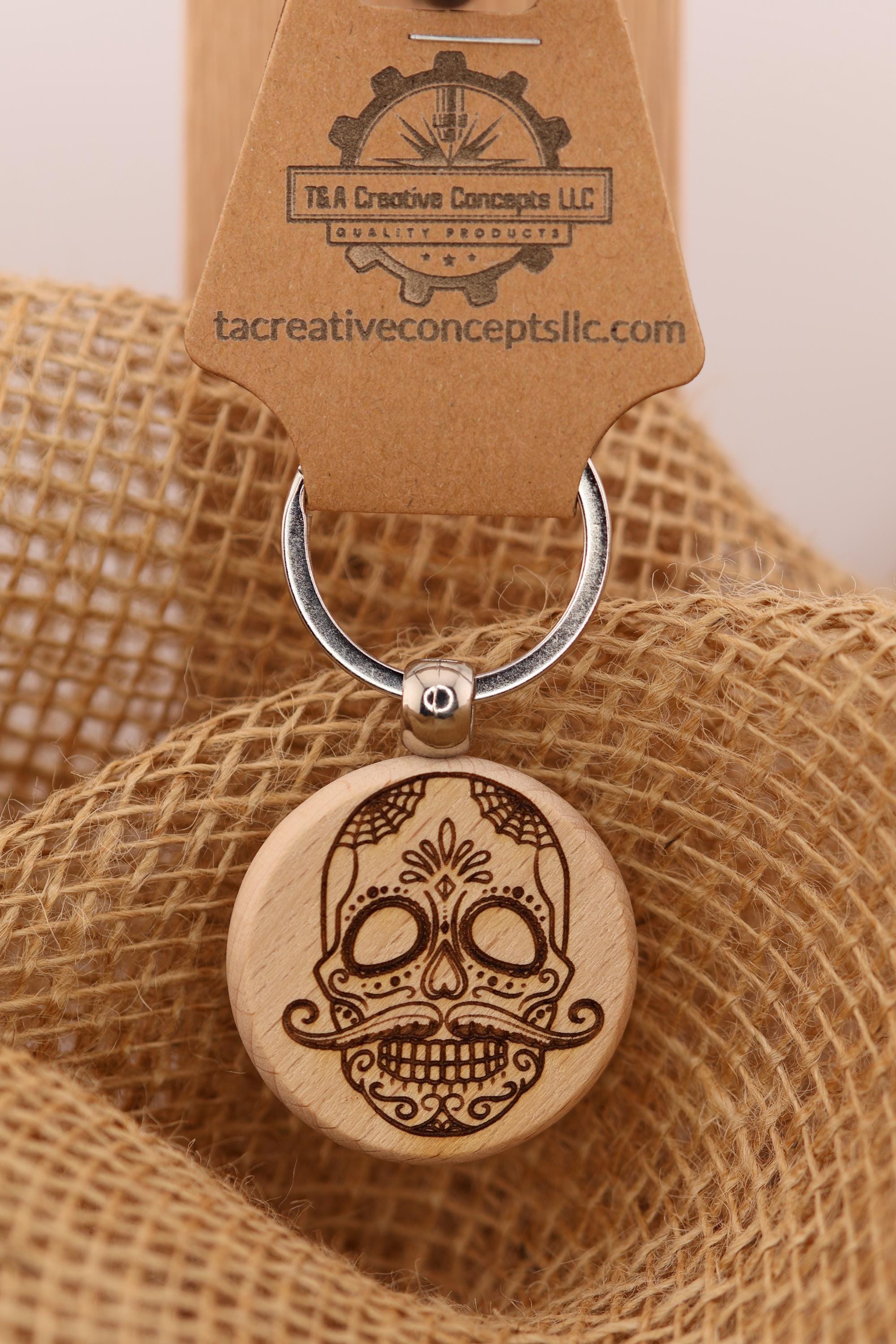 May include: A wooden keychain featuring a detailed skull design. The skull has intricate etched details, including a mustache, and is attached to a silver key ring. The keychain is branded with 'T & A Creative Concepts LLC' and the website 'tacreativeconceptsllc.com'.