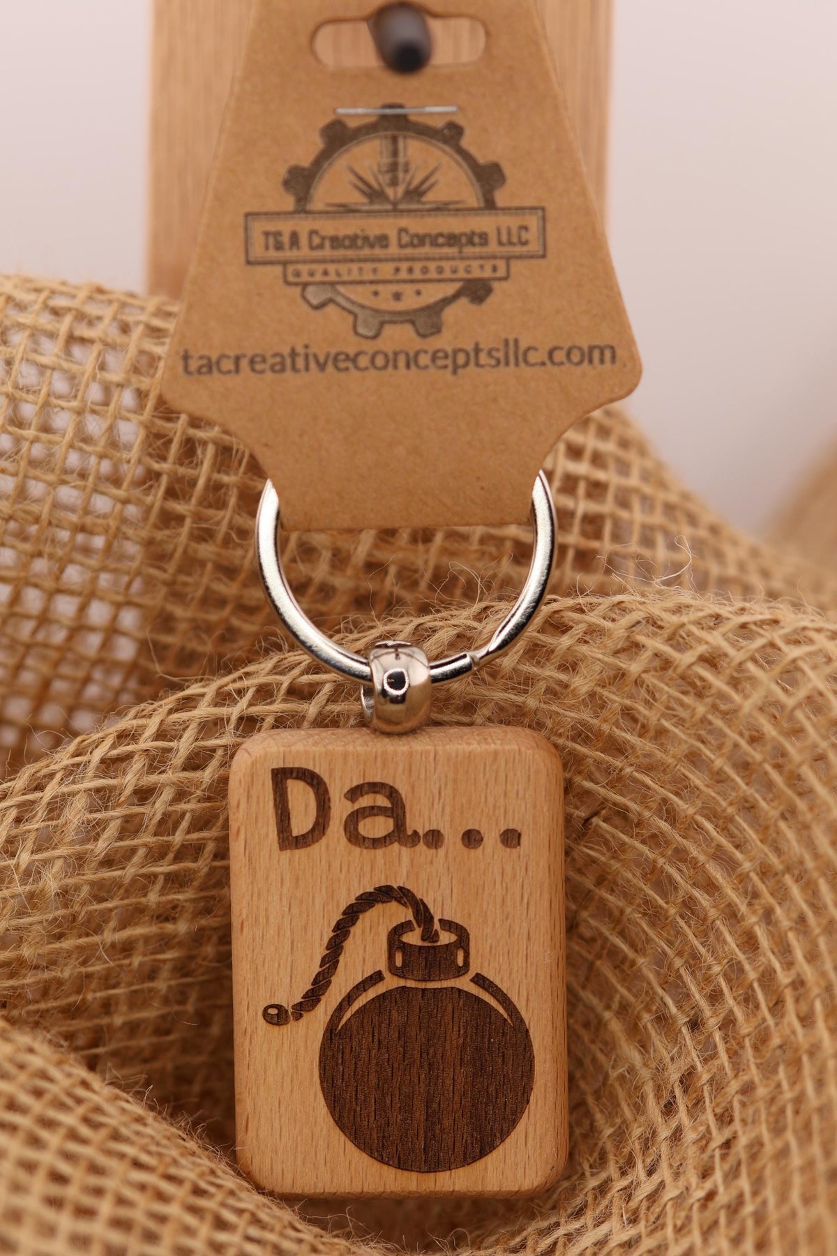 May include: A wooden keychain with a bomb design. The keychain is rectangular with rounded corners and a silver key ring. The bomb is brown with a rope and fuse design, and the word 'Da...' is engraved above it. The keychain is attached to a brown cardboard tag with the text 'T&A Creative Concepts LLC' and a website address.