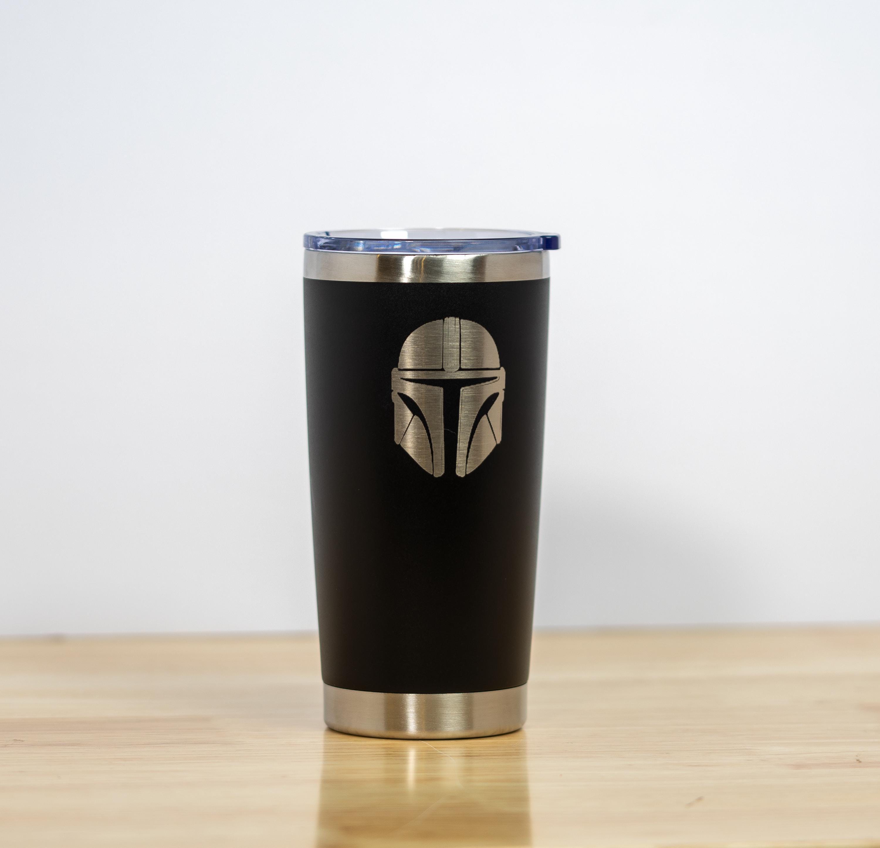 May include: A black stainless steel tumbler with a silver rim and lid. The tumbler features a silver Mandalorian helmet design. The tumbler is designed to keep beverages hot or cold.