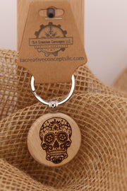 May include: A wooden keychain with a sugar skull design. The sugar skull is engraved in brown on a round, light-colored wooden pendant. The keychain has a silver ring and a brown tag with the text 'T&A Creative Concepts LLC' and 'tacreativeconceptsllc.com'.