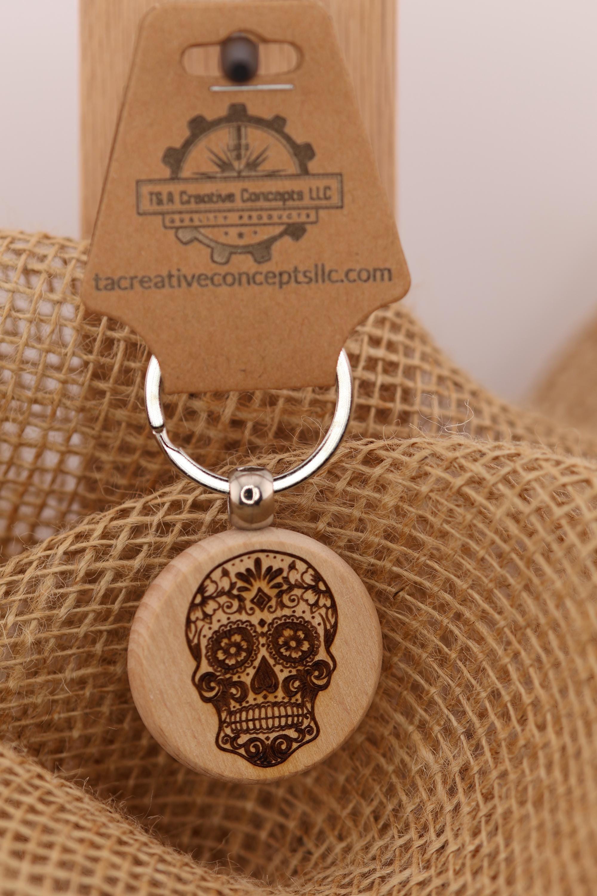 May include: A wooden keychain with a sugar skull design. The sugar skull is engraved in brown on a round, light-colored wooden pendant. The keychain has a silver ring and a brown tag with the text 'T&A Creative Concepts LLC' and 'tacreativeconceptsllc.com'.
