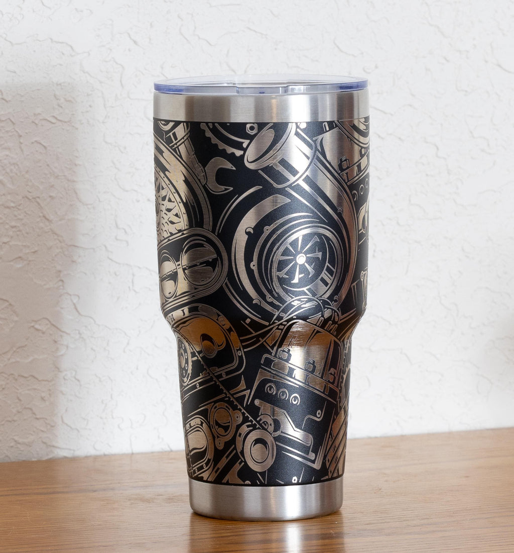May include: Stainless steel tumbler with a black background and a detailed silver-colored engine parts design. The tumbler has a clear lid and a silver base and rim. The design includes gears, turbochargers, and other mechanical components.