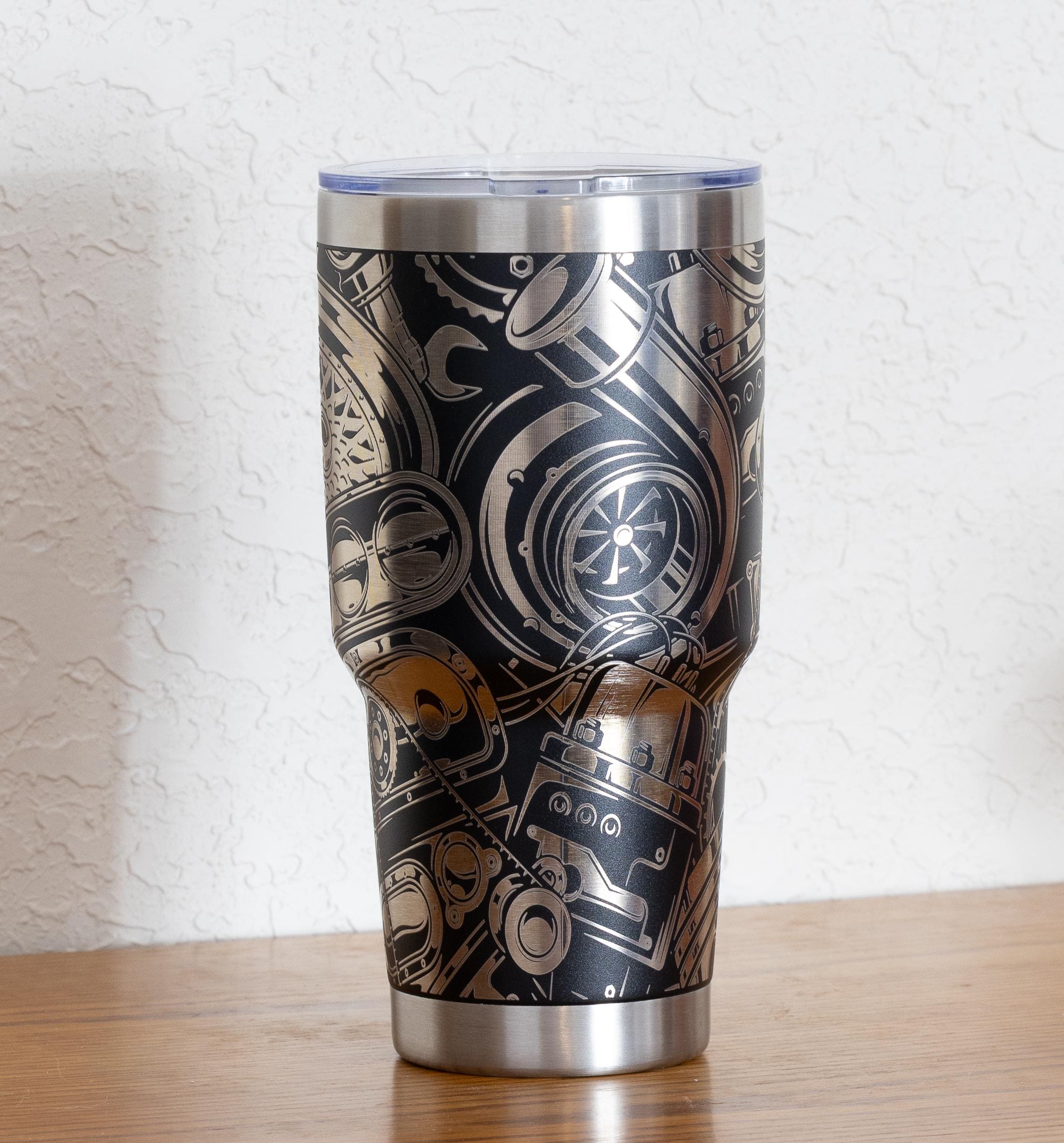 May include: Stainless steel tumbler with a black background and a detailed silver-colored engine parts design. The tumbler has a clear lid and a silver base and rim. The design includes gears, turbochargers, and other mechanical components.