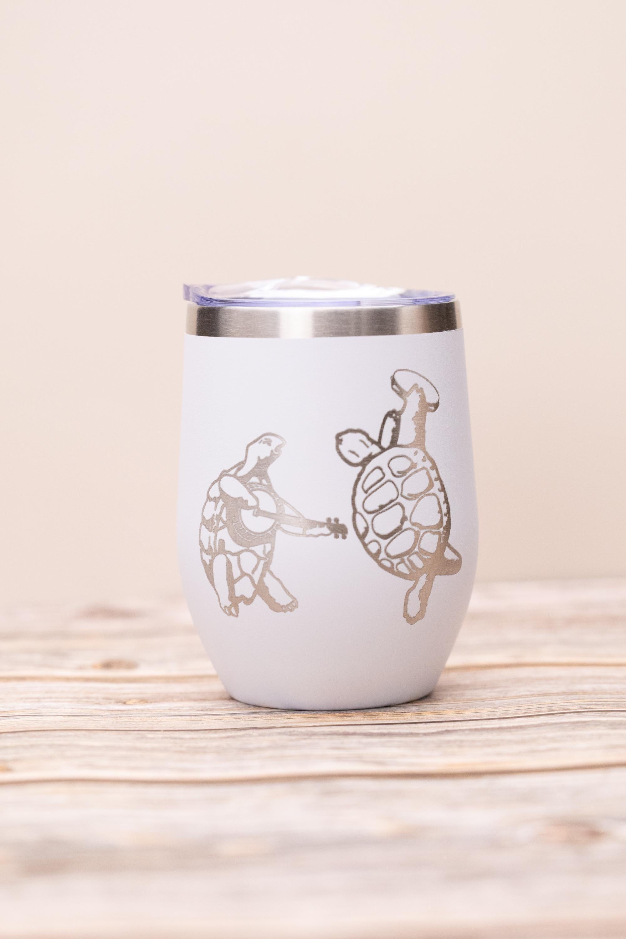 May include: White insulated wine tumbler with a silver rim and lid. The tumbler features two silver illustrations of turtles, one playing a guitar and the other dancing. The design is whimsical and fun.