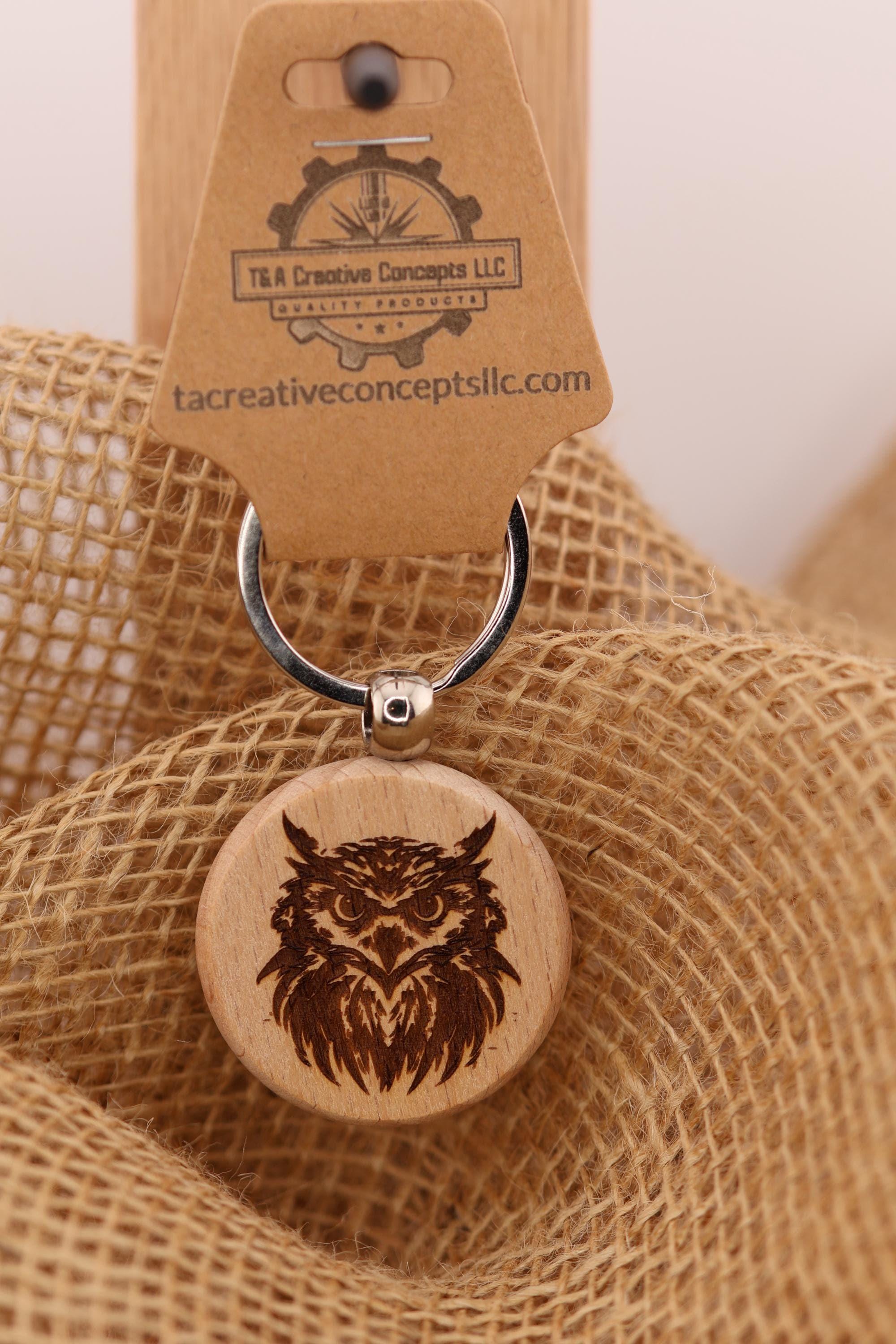 May include: A wooden keychain with a laser-engraved owl design. The owl is intricately detailed with feathers and facial features. The keychain is attached to a silver metal ring and a brown cardboard tag with the text 'T&A Creative Concepts LLC QUALITY PRODUCTS' and 'tacreativeconceptsllc.com'.