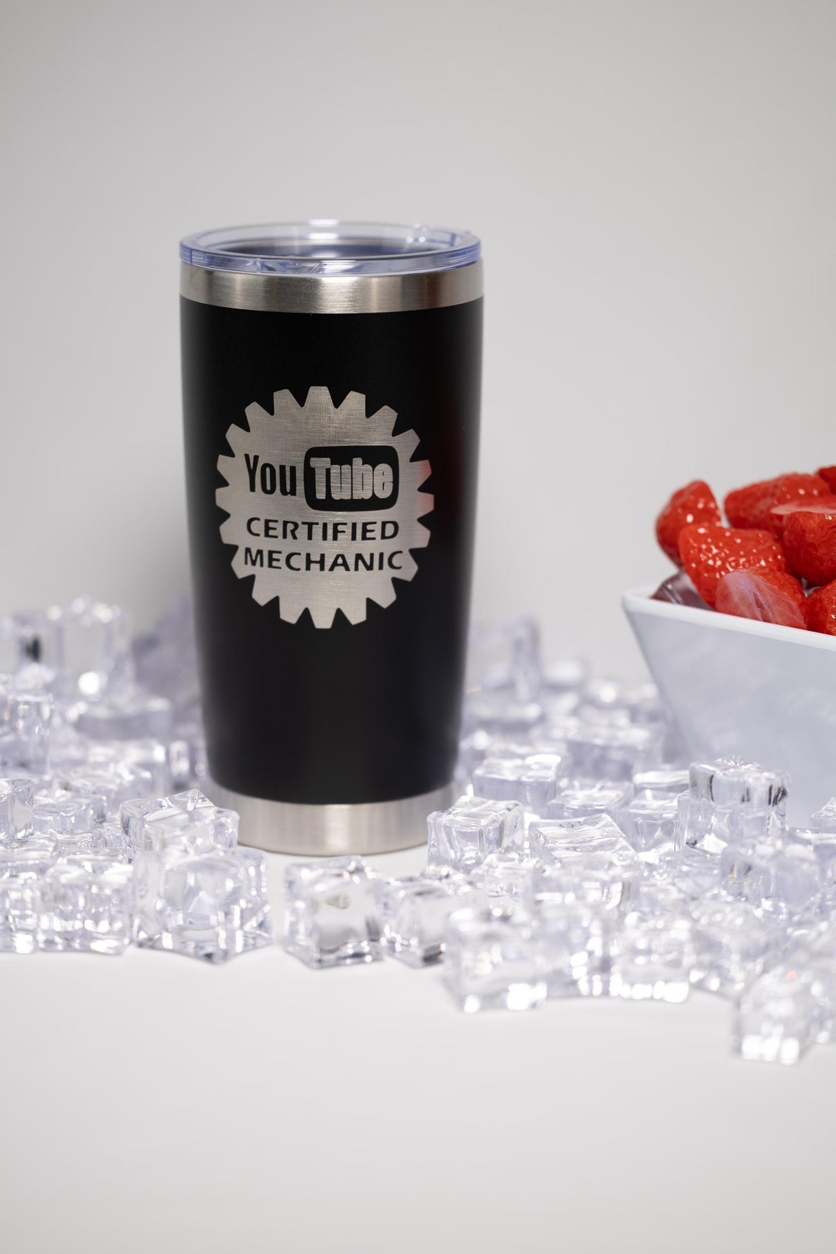 A black tumbler with a silver lid, featuring a logo that says "YouTube Certified Mechanic" on it, placed on a bed of small ice cubes.