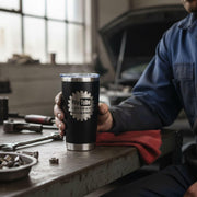 A person is sitting at a workbench in a garage, holding a black tumbler with the YouTube logo and the text "Certified Mechanic" printed on it.