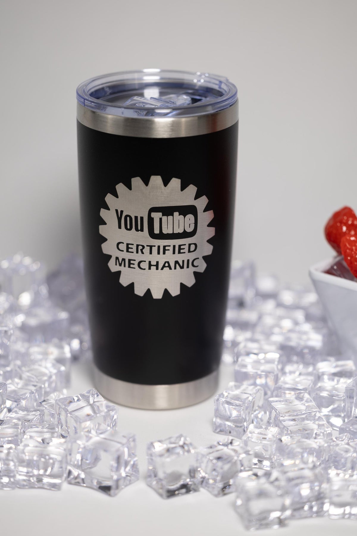 A black tumbler with a silver lid, featuring the text "YouTube Certified Mechanic" printed on it, surrounded by ice cubes and a bowl of strawberries.