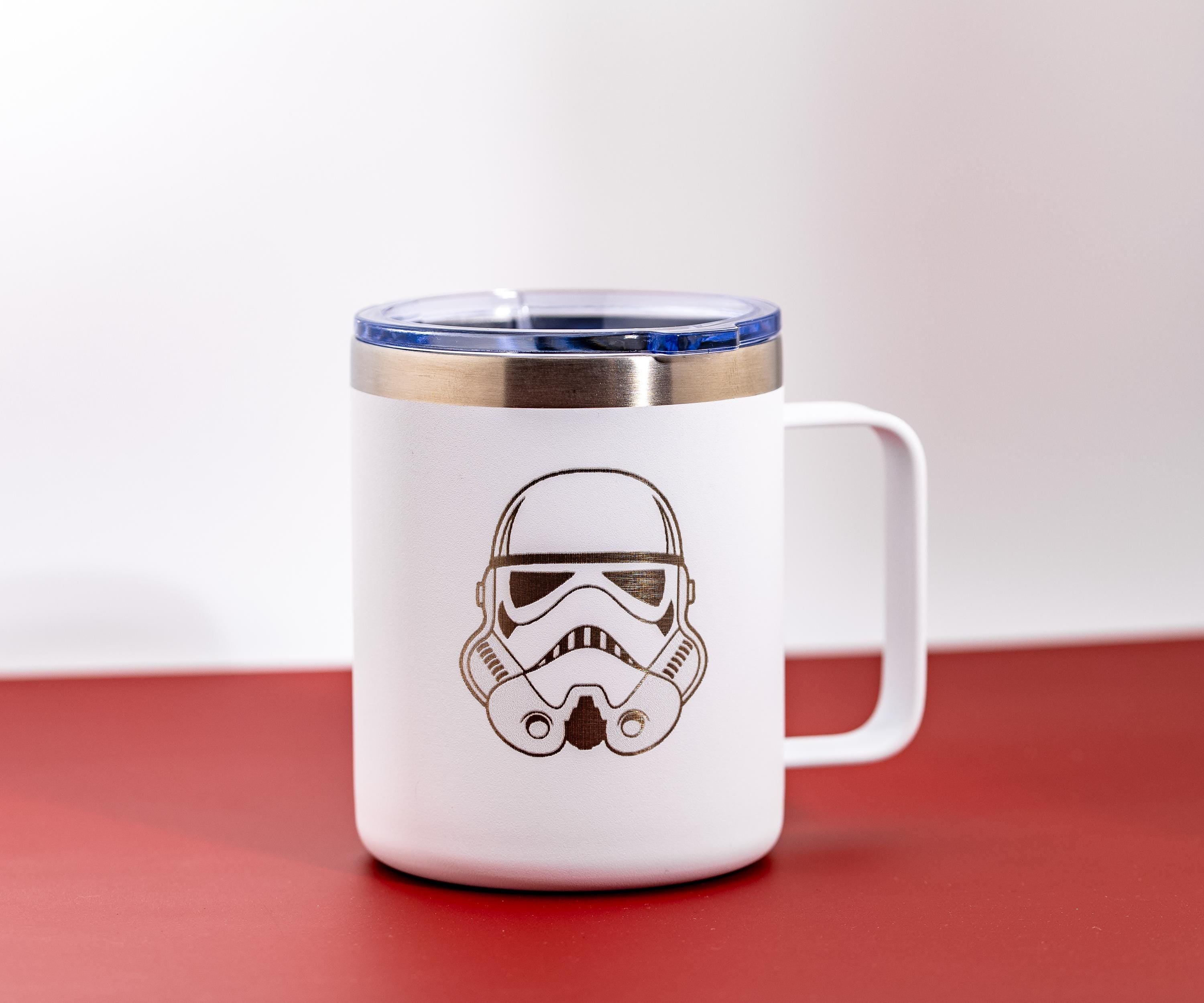 May include: White travel mug with a silver rim and a blue lid. The mug features a black outline of a Stormtrooper helmet. The mug has a white handle and is on a red surface.