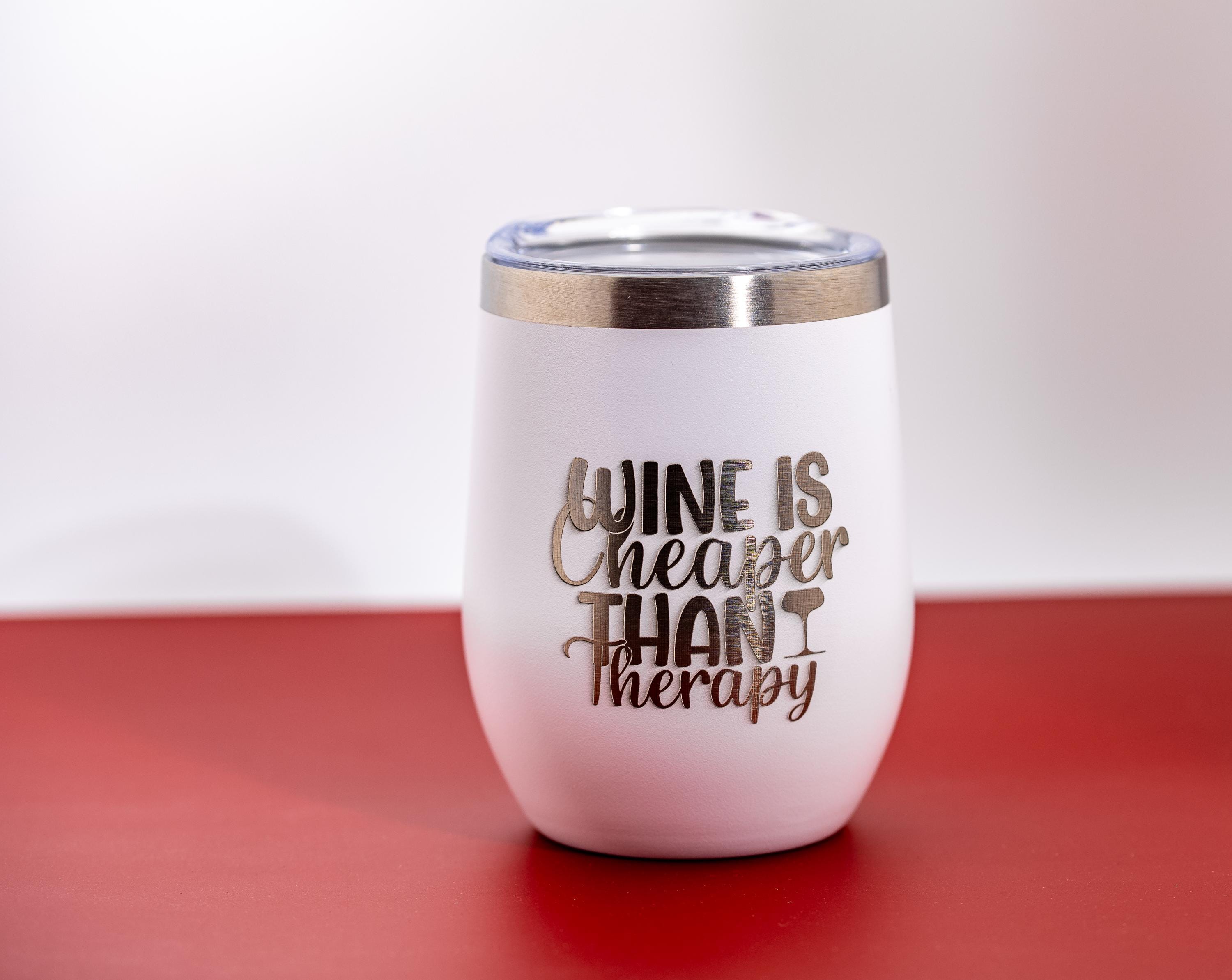 May include: White insulated wine tumbler with a silver rim and clear lid. The tumbler has the phrase "WINE IS CHEAPER THAN THERAPY" in silver lettering. The tumbler sits on a red surface against a white background.