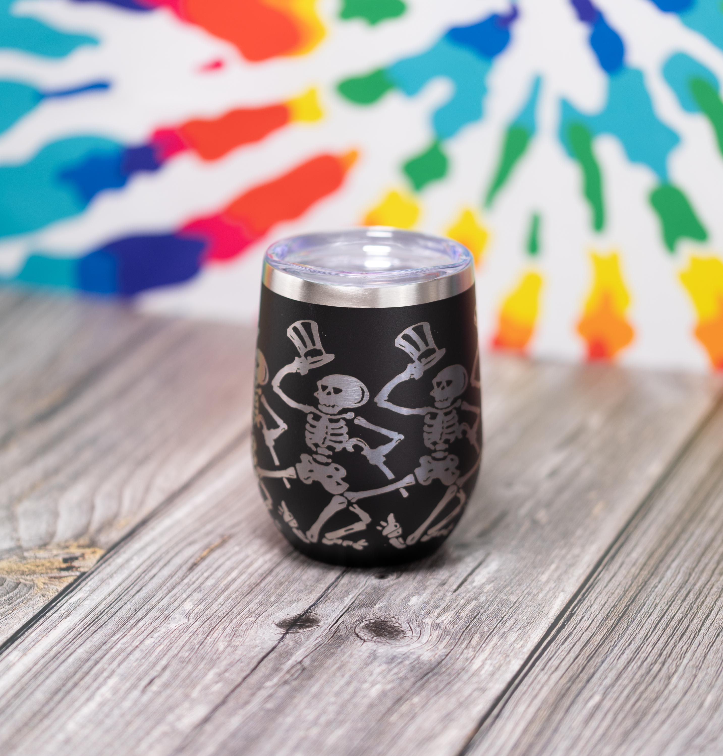 May include: Black stemless wine tumbler with a silver rim and lid. The tumbler features a silver illustration of dancing skeletons wearing top hats. The background is a tie-dye pattern in multiple colors.