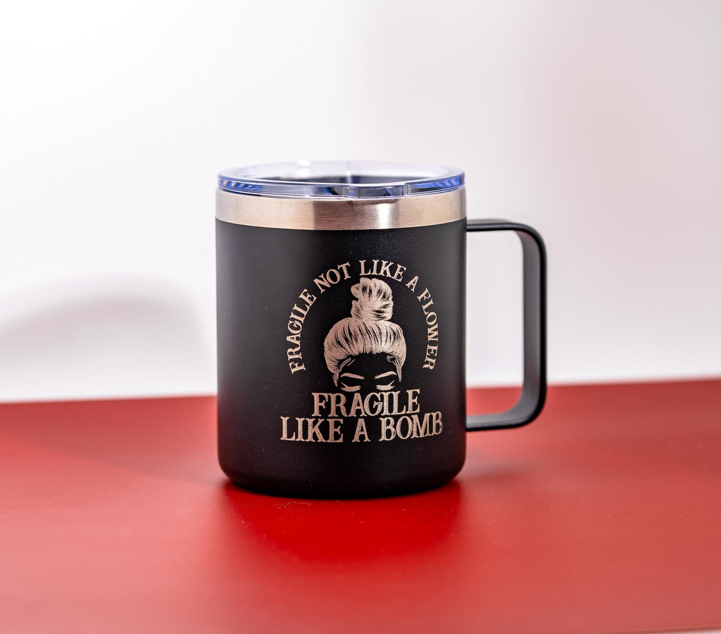 May include: Black travel mug with a silver rim and handle. The mug features the text "FRAGILE NOT LIKE A FLOWER FRAGILE LIKE A BOMB" around a woman's face. The mug has a black handle and a clear lid.