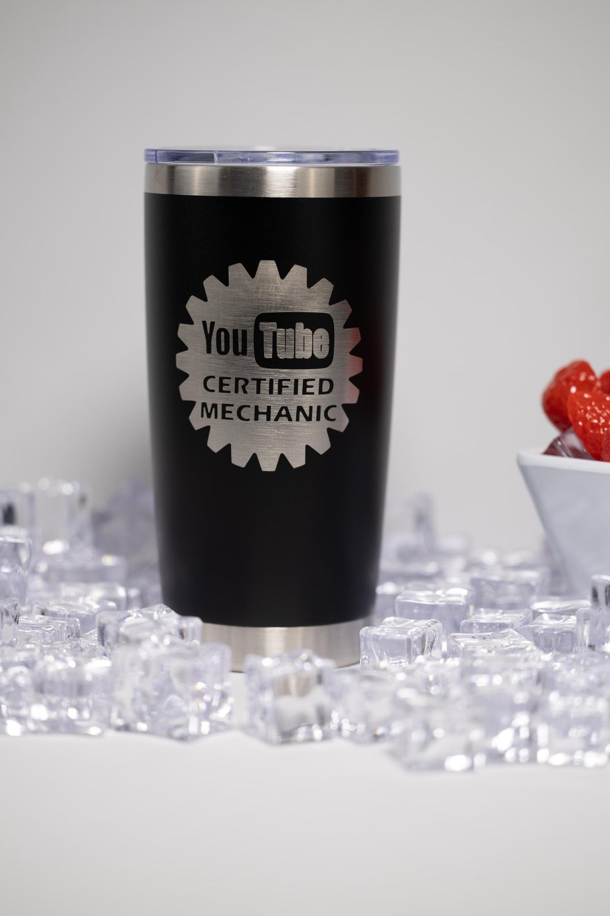 A black tumbler with a silver lid, featuring a logo that says "YouTube Certified Mechanic" on it, placed on a surface with ice cubes.