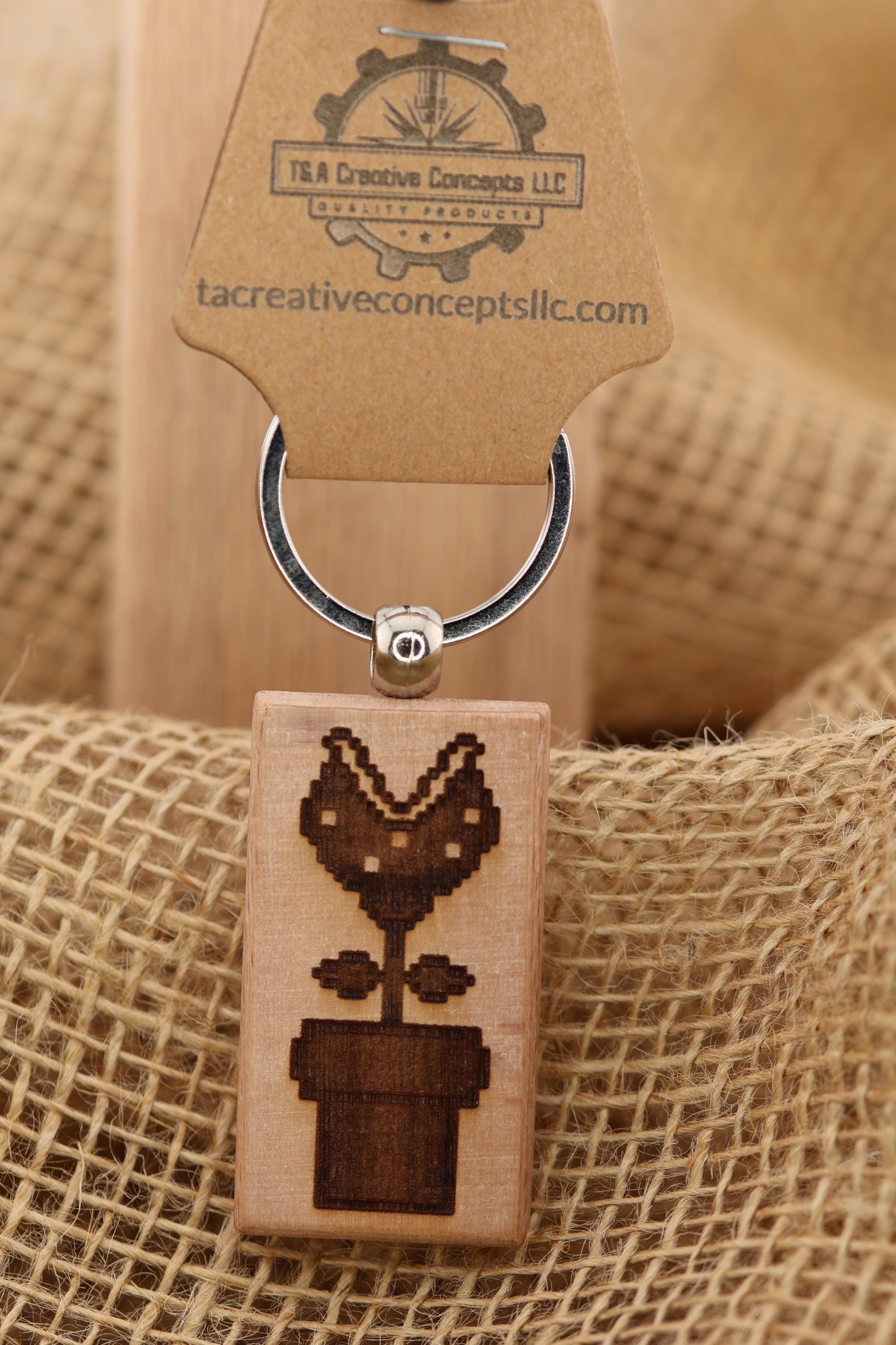 May include: A wooden keychain featuring a pixelated design of a Piranha Plant in a pot. The plant has a brown pot with a green stem and a brown flower with a mouth. The keychain has a silver ring and a brown tag with the text 'T&A Creative Concepts LLC Quality Products' and the website 'tacreativeconceptsllc.com'.