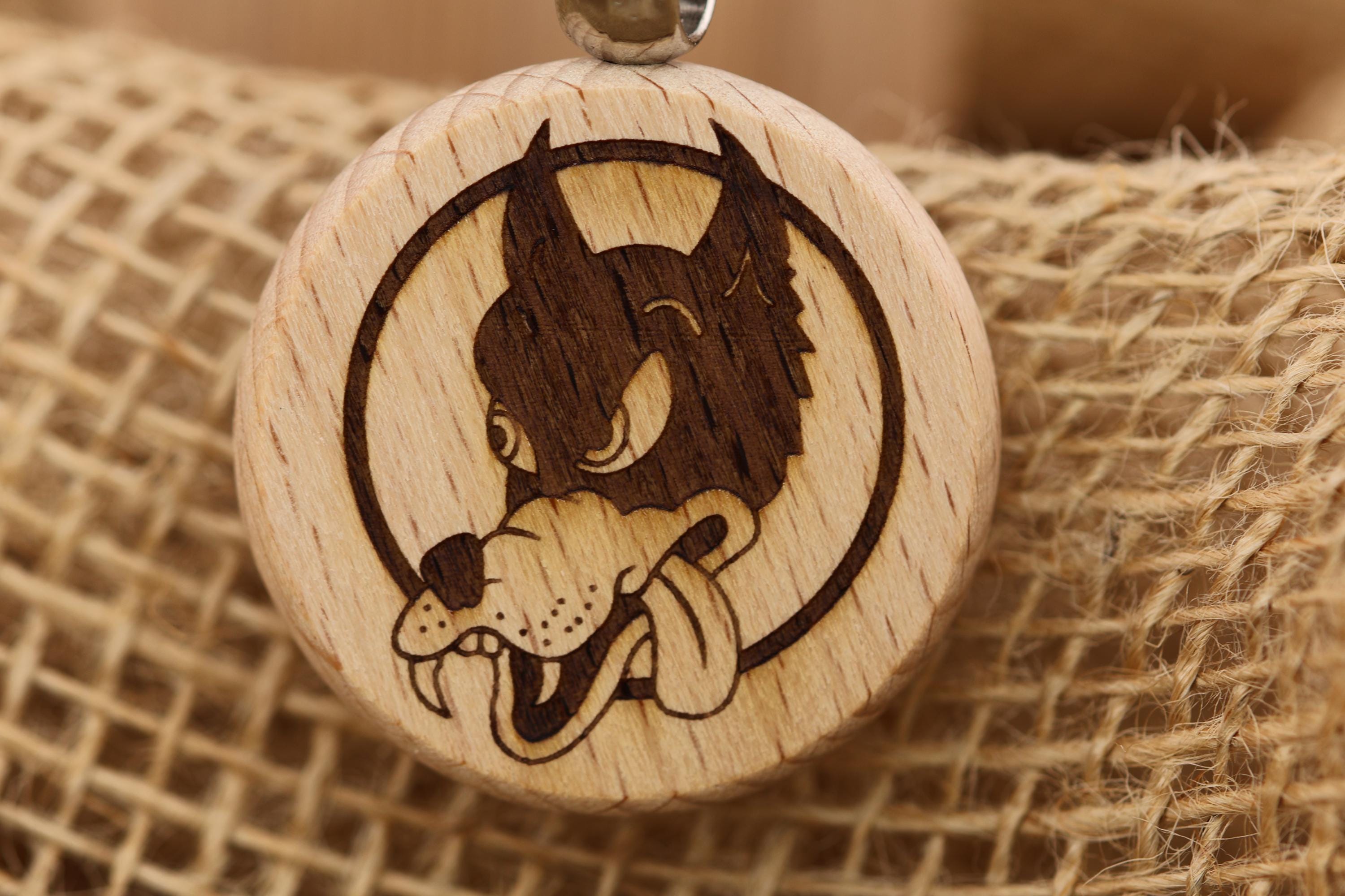May include: A round wooden pendant with a wolf design. The wolf is dark brown and inside a black circle. The wolf has a tongue sticking out. The pendant is light brown and has a silver loop at the top for a necklace.