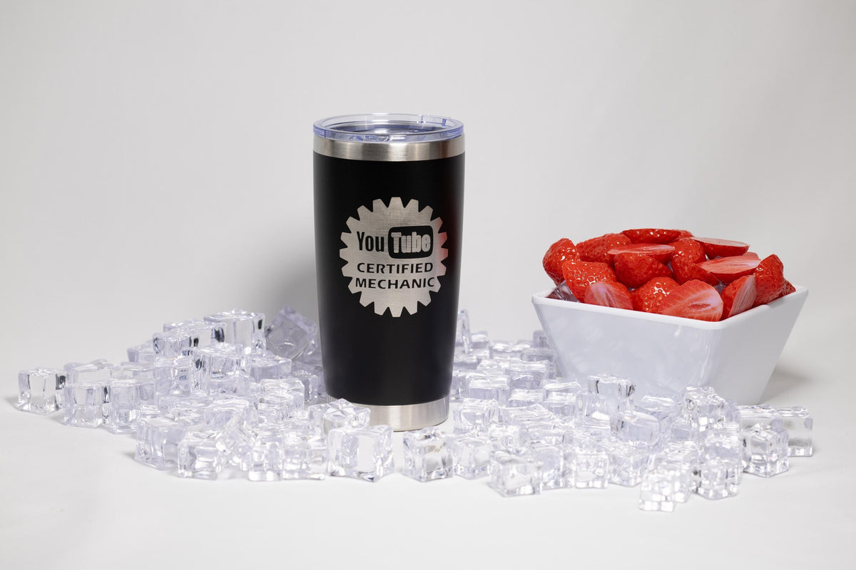 A black tumbler with a white logo and text, a white bowl filled with red strawberries, and a pile of clear ice cubes on a white background.