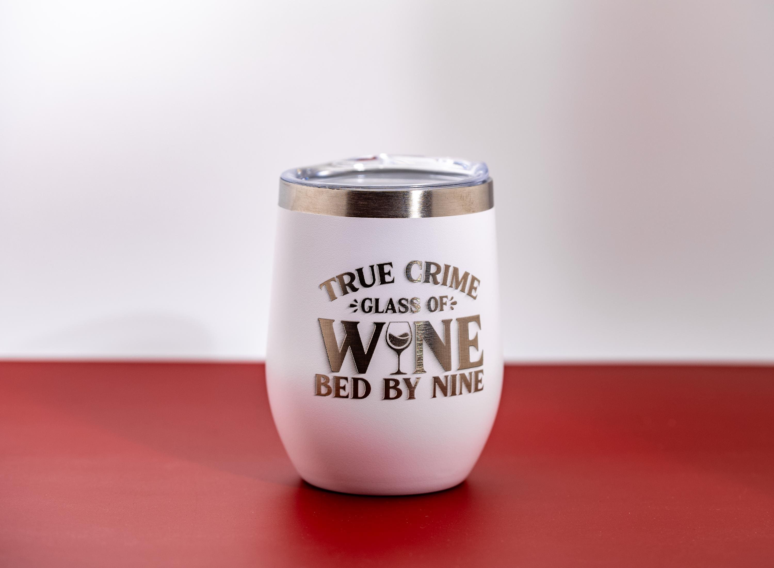 May include: White insulated wine tumbler with a silver rim and a clear lid. The tumbler features the text "TRUE CRIME GLASS OF WINE BED BY NINE" in silver lettering. The tumbler is on a red surface.