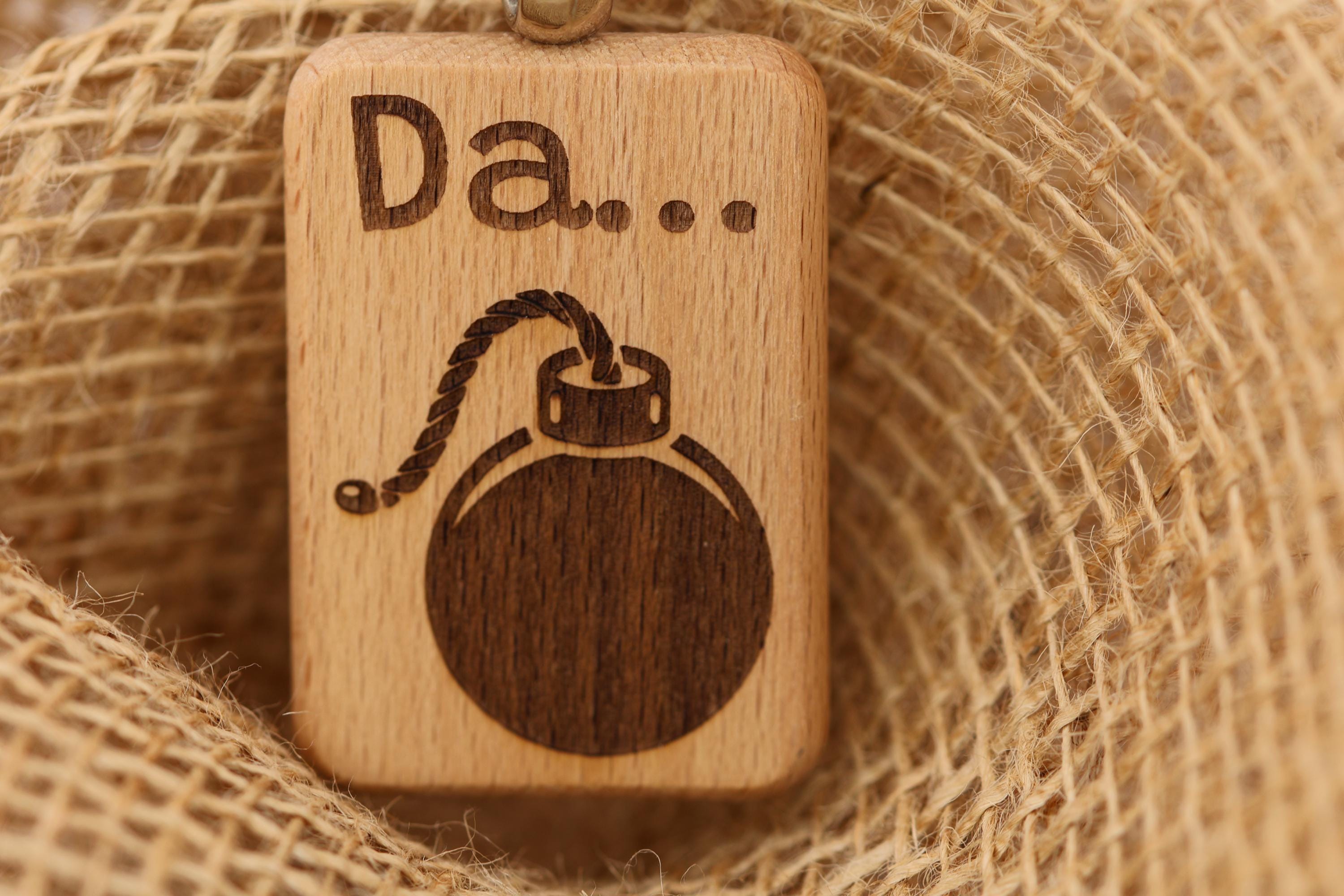 May include: A rectangular wooden keychain with the word 'Da...' engraved at the top. Below the text is a bomb illustration with a rope fuse. The bomb is a dark brown color, and the keychain is light brown. The background is a textured, tan fabric.