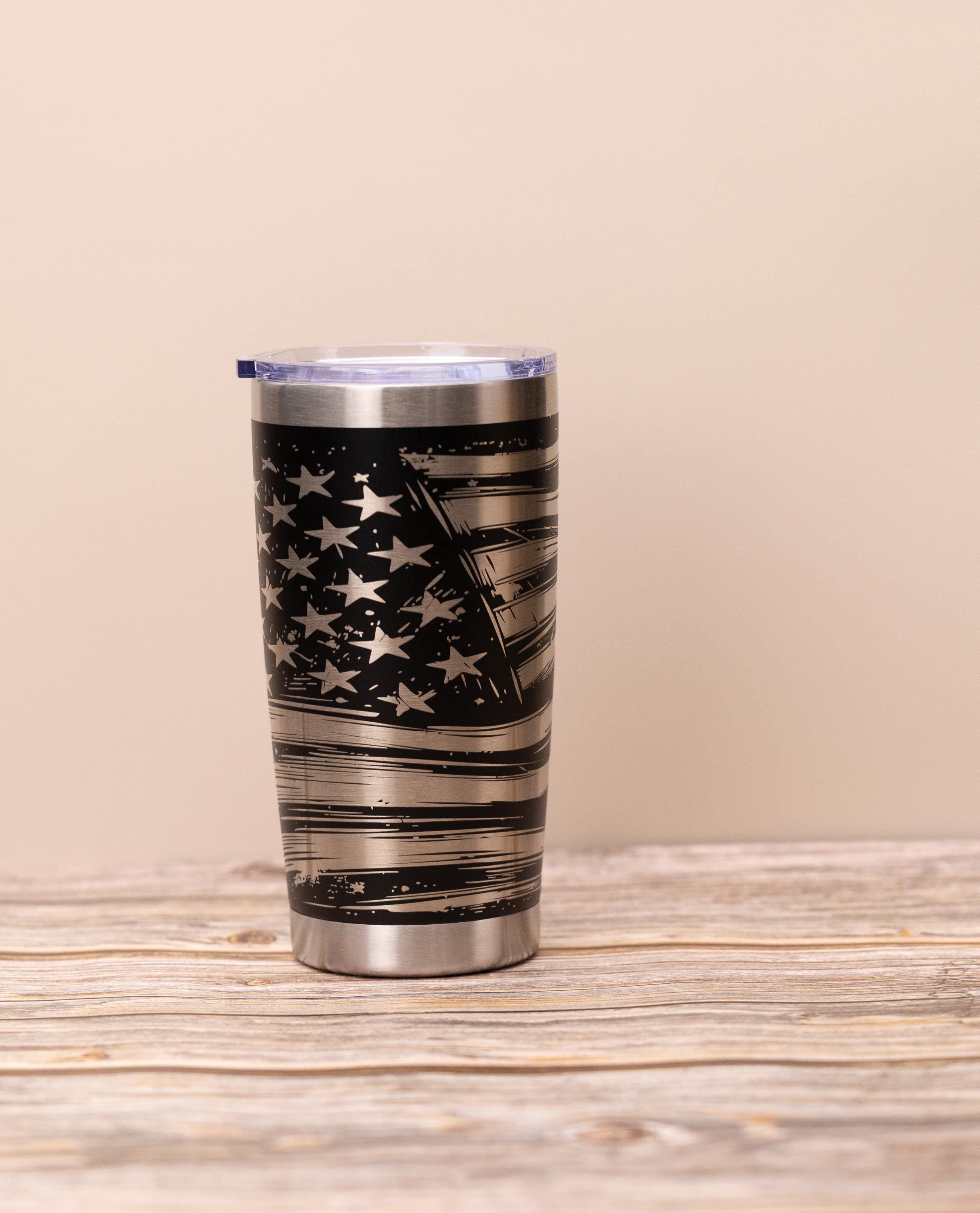 May include: Stainless steel tumbler with a black and silver American flag design. The flag features stars and stripes with a distressed, brushed metal effect. The tumbler has a clear lid and is designed for beverages.