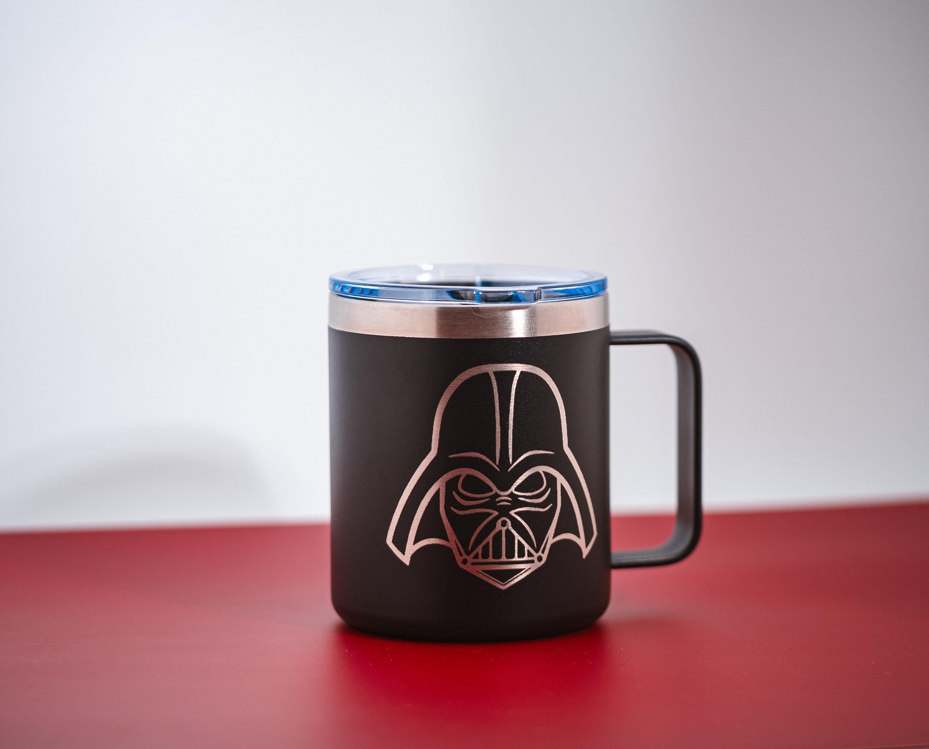 May include: Black travel mug with a silver rim and handle, featuring a rose gold illustration of Darth Vader's helmet. The mug has a blue lid and sits on a red surface. A Star Wars themed product.