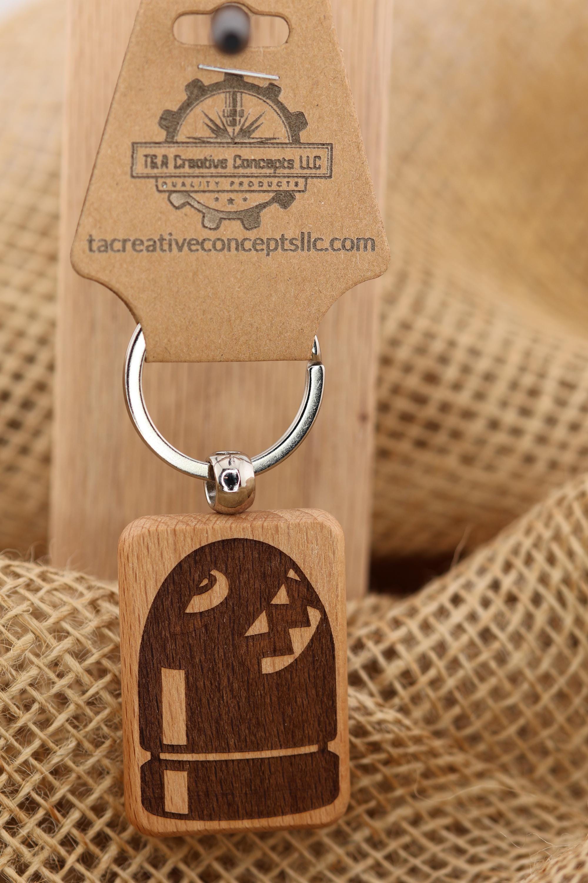May include: A wooden keychain with a dark brown engraved design. The keychain features a rectangular wooden block with a silver key ring and a tag. The tag has the text 'T&R Creative Concepts LLC' and 'tacreativeconceptsllc.com'. The engraved design on the block is a cartoon character.