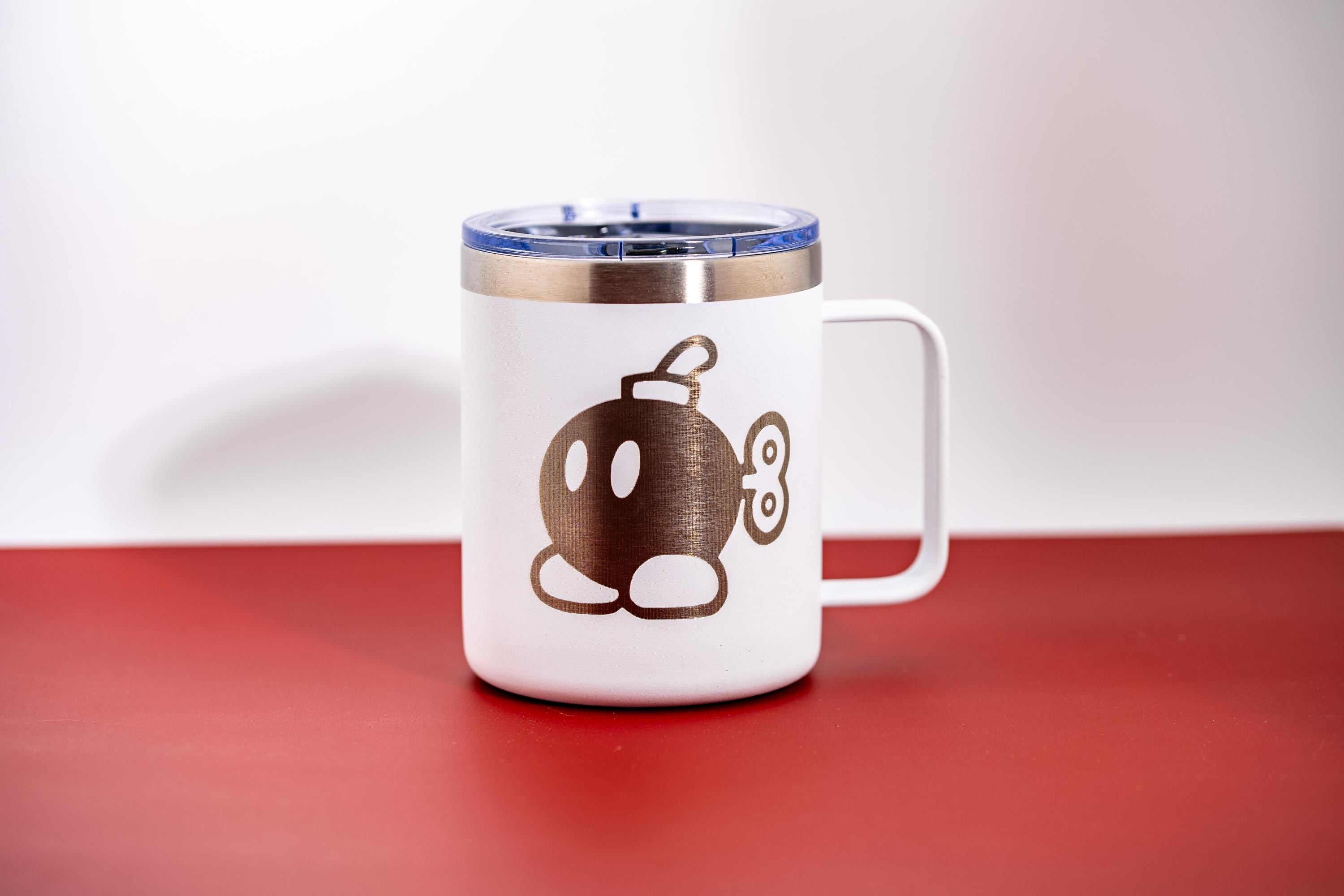 May include: White travel mug with a blue lid and a handle. The mug features a brown cartoon bomb character design. The mug is on a red surface with a white background.